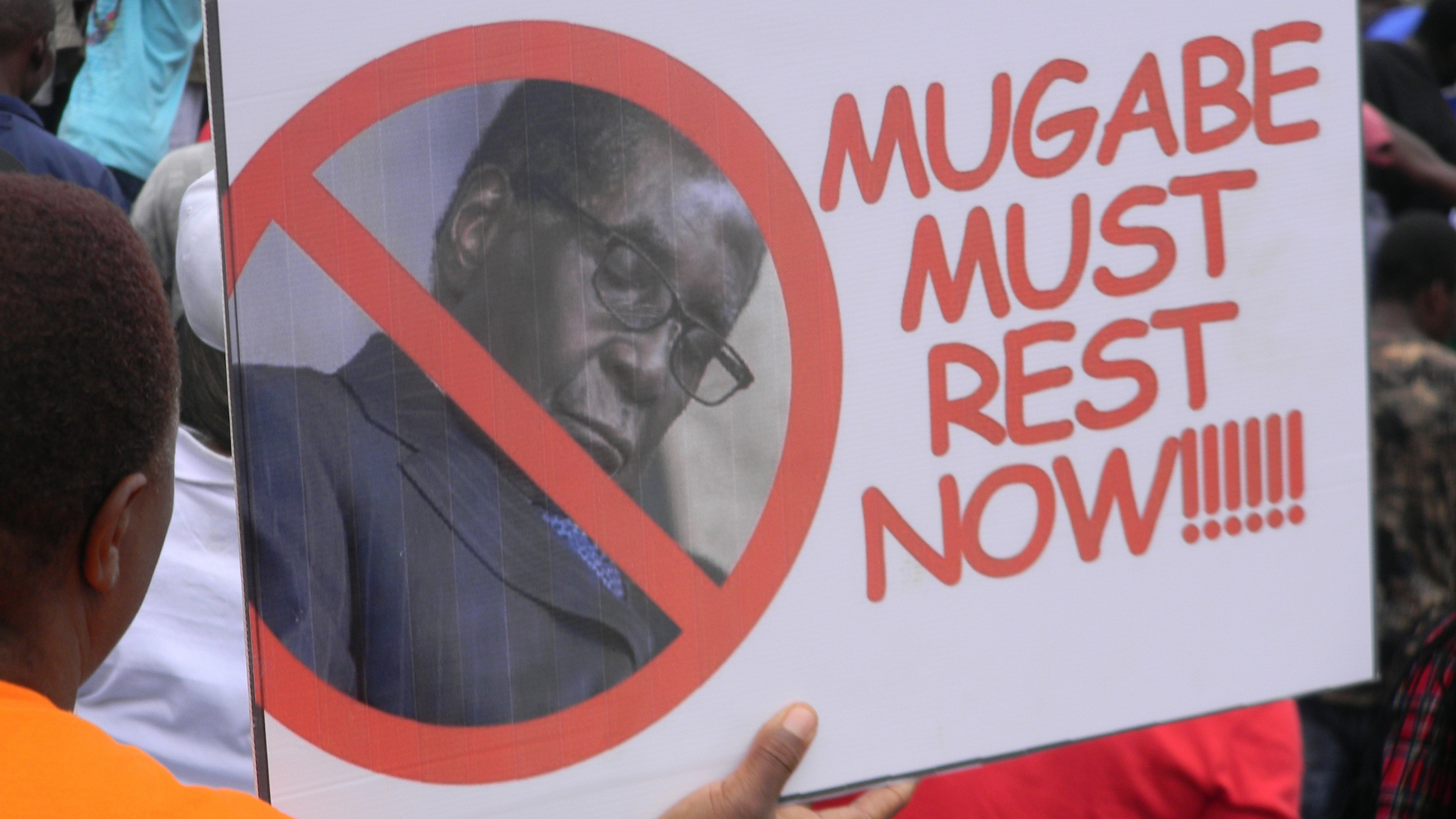 Cette image montre une personne tenant une pancarte lors d'une manifestation. La pancarte arbore un grand symbole barré, avec une photo d'un homme, généralement associé à un discours politique. Le texte sur la pancarte se lit : "MUGABE MUST REST NOW", exprimant un message clair de résistance ou de demande de cessation à l'égard de cette personne. En arrière-plan, on peut voir une foule, suggérant un rassemblement où des gens partagent des opinions fortes sur un sujet d'actualité. L'ambiance générale semble être celle d'une protestation.