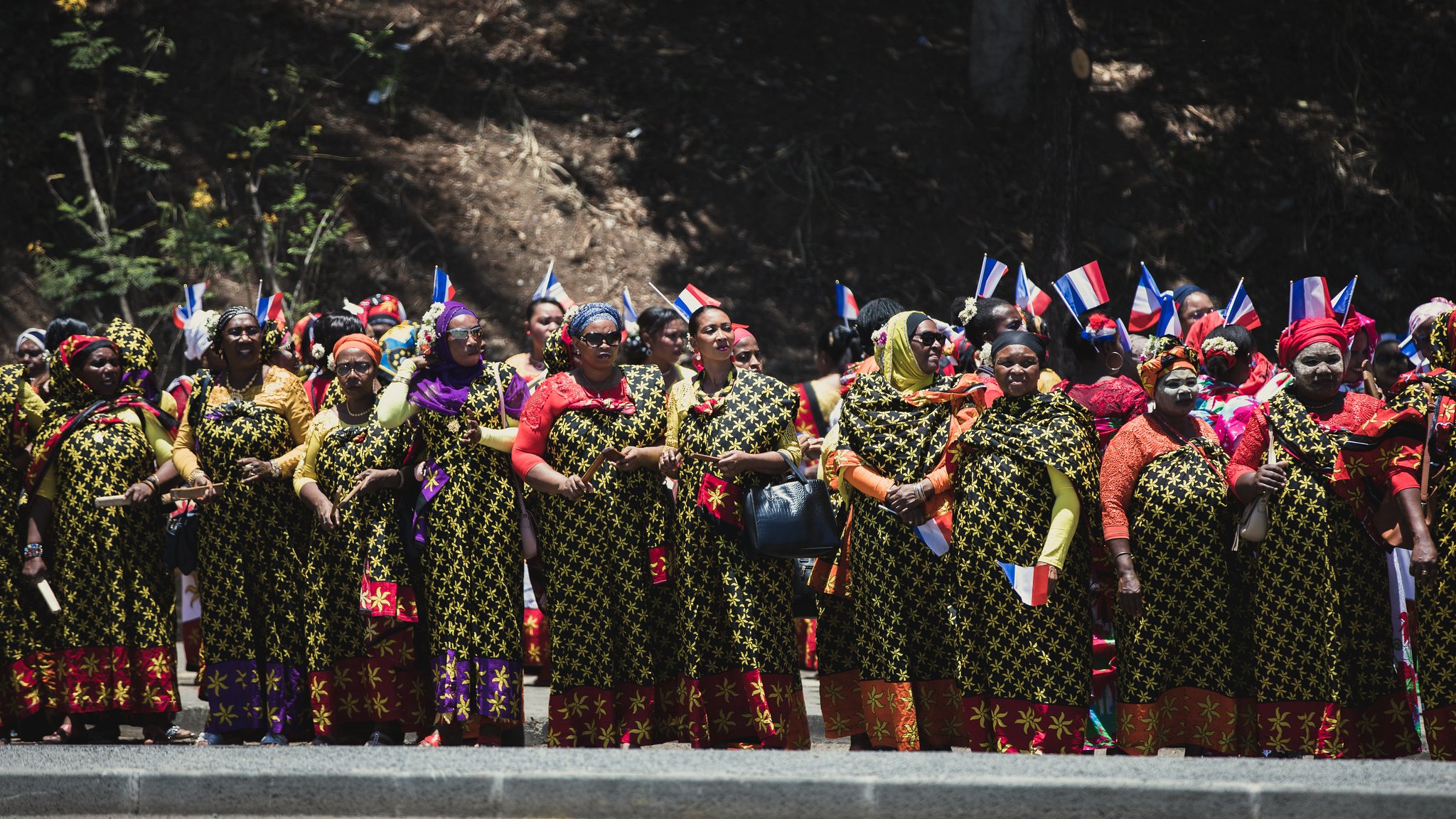 L'image montre un groupe de femmes réunies en ligne, vêtues de robes longues aux motifs floraux colorés. Chaque femme porte des accessoires traditionnels, comme des foulards vifs. Certaines tiennent des drapeaux aux couleurs bleu, blanc et rouge, symbolisant leur fierté nationale. L'arrière-plan est naturel, avec des arbres, et l'ambiance semble festive et engagée, suggérant une célébration ou un événement culturel. Les femmes affichent des expressions de joie et d'unité.