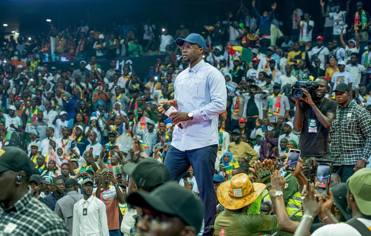 L'image représente un événement animé dans une grande salle remplie de personnes. Au premier plan, un homme en tenue décontractée, portant une chemise claire et une casquette, marche avec assurance sur la scène. Il semble interagir avec le public, qui se compose de nombreux spectateurs enthousiastes, certains levant les mains en signe d'encouragement. La foule est colorée, avec des personnes habillées en diverses tenues, et des drapeaux ou accessoires visibles, suggérant un fort esprit communautaire. En arrière-plan, on peut voir des caméras et des membres de l'équipe de sécurité, ce qui indique que l'événement est de grande envergure. L'atmosphère est d'excitation et d'engagement collectif.
