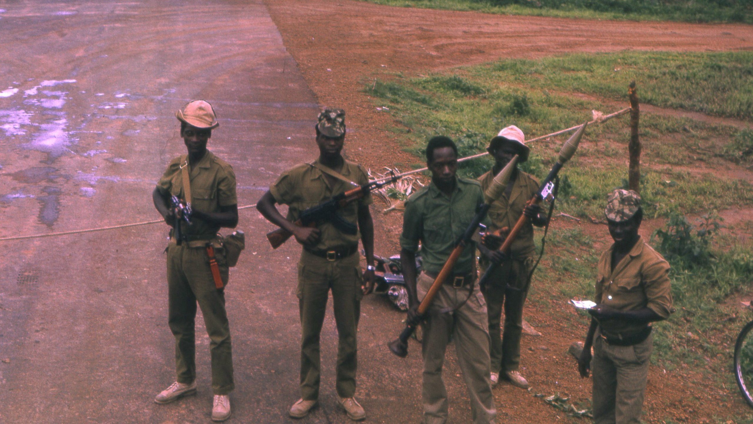 L'image montre un groupe de cinq soldats en uniforme, debout sur une route non pavée. Ils portent des chapeaux, et chacun d'eux tient une arme à feu. Le sol est poussiéreux, avec quelques zones humides visibles. En arrière-plan, on voit une végétation luxuriante, typique d'un environnement tropical. Les soldats affichent des postures sérieuses, et certains d'entre eux paraissent concentrés sur leur environnement. Cette scène suggère un contexte de sécurité ou de patrouille.
