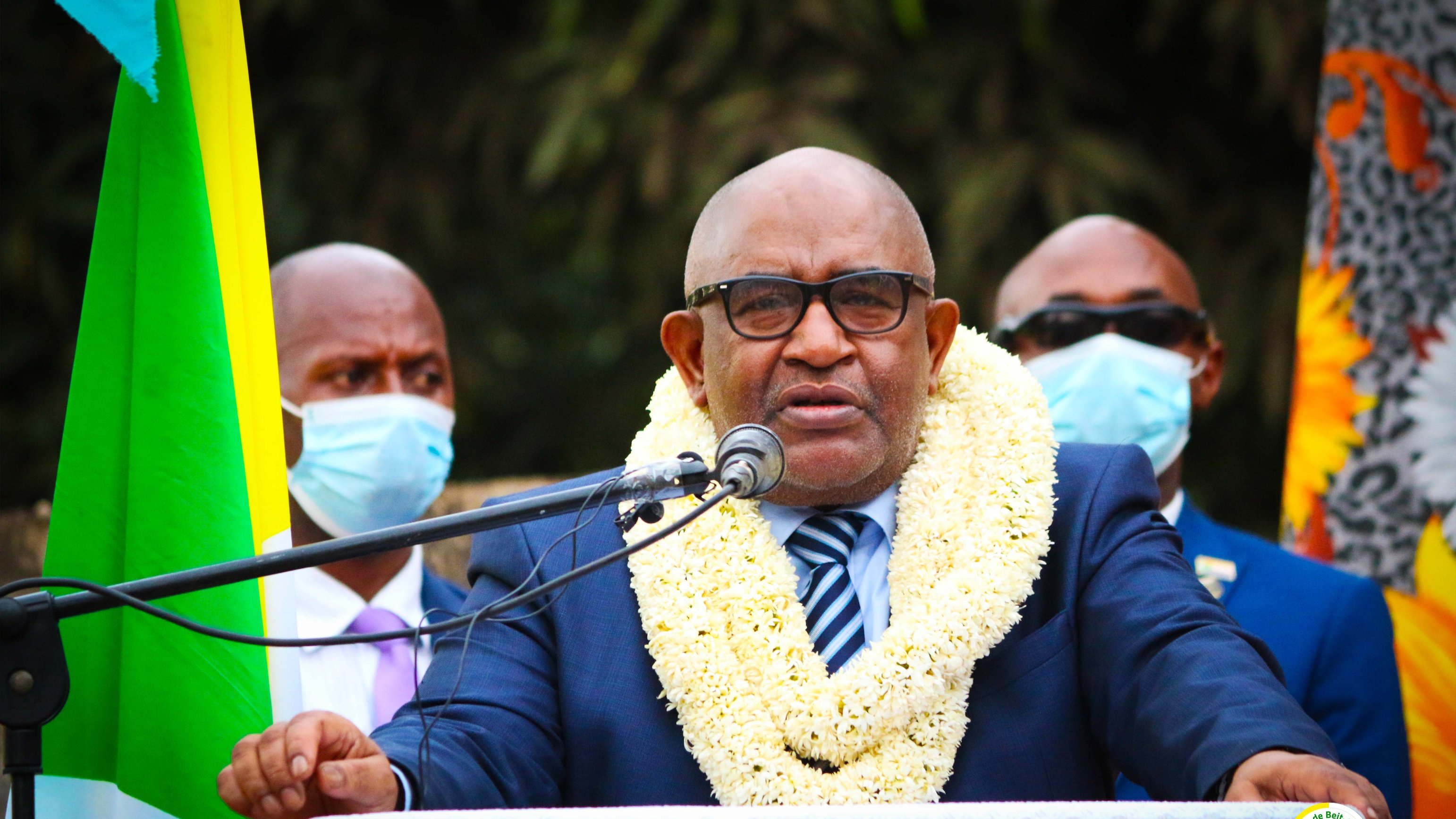 Sur cette image, un homme est debout devant un microphone, en train de s'exprimer. Il porte un costume sombre avec une chemise claire et une cravate rayée. Autour de son cou, une couronne de fleurs blanches et jaunes symbolise un accueil ou un hommage. En arrière-plan, plusieurs personnes l'écoutent attentivement, certaines portant des masques de protection. À gauche, on peut voir des drapeaux aux couleurs vives qui ajoutent une touche festive à la scène. L'ambiance semble sérieuse et solennelle.