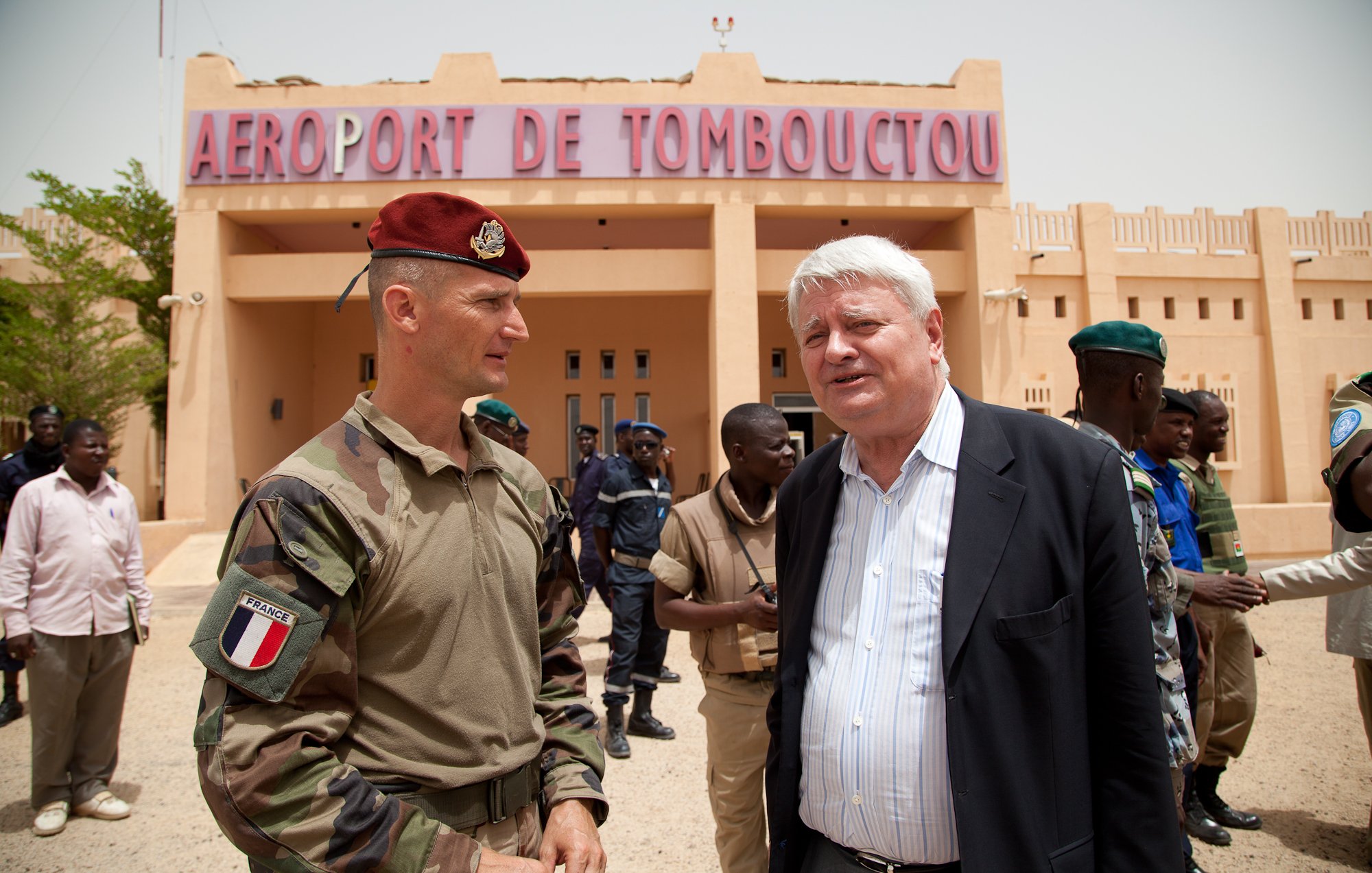 L'image montre une scène à l'aéroport de Tombouctou, avec un bâtiment beige en arrière-plan portant le nom "AÉROPORT DE TOMBOUCTOU" en lettres rouges. Au premier plan, deux hommes sont en conversation. L'un est en uniforme militaire, portant un béret rouge, tandis que l'autre est habillé en costume. Autour d'eux, plusieurs personnes en uniforme, probablement des militaires et des policiers, sont présentes, créant une atmosphère chargée d'activités. Le ciel est clair et le paysage indique une région chaude, avec un sol désertique.