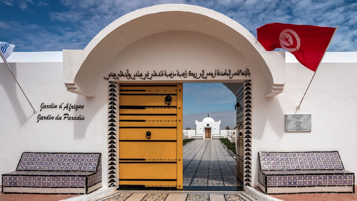 Imaginez un lieu paisible et accueillant, avec une grande porte jaune ornée de motifs géométriques. Cette porte s'ouvre sur un jardin magnifique, symbolisant la beauté et la diversité de la nature. Deux bancs placés de chaque côté de l'entrée, recouverts de carrelage coloré, invitent à la détente. Au-dessus de la porte, une inscription en arabe et en français nous invite à découvrir ce jardin, intitulé "Jardin d'Afrique, Jardin du Paradis". De chaque côté, un drapeau flotte, ajoutant une touche de couleur vive à la scène. Le ciel est lumineux et parsemé de nuages, apportant une lumière douce qui souligne la sérénité de cet endroit. L'ambiance générale est calme et harmonieuse, évoquant un espace de ressourcement et de contemplation.