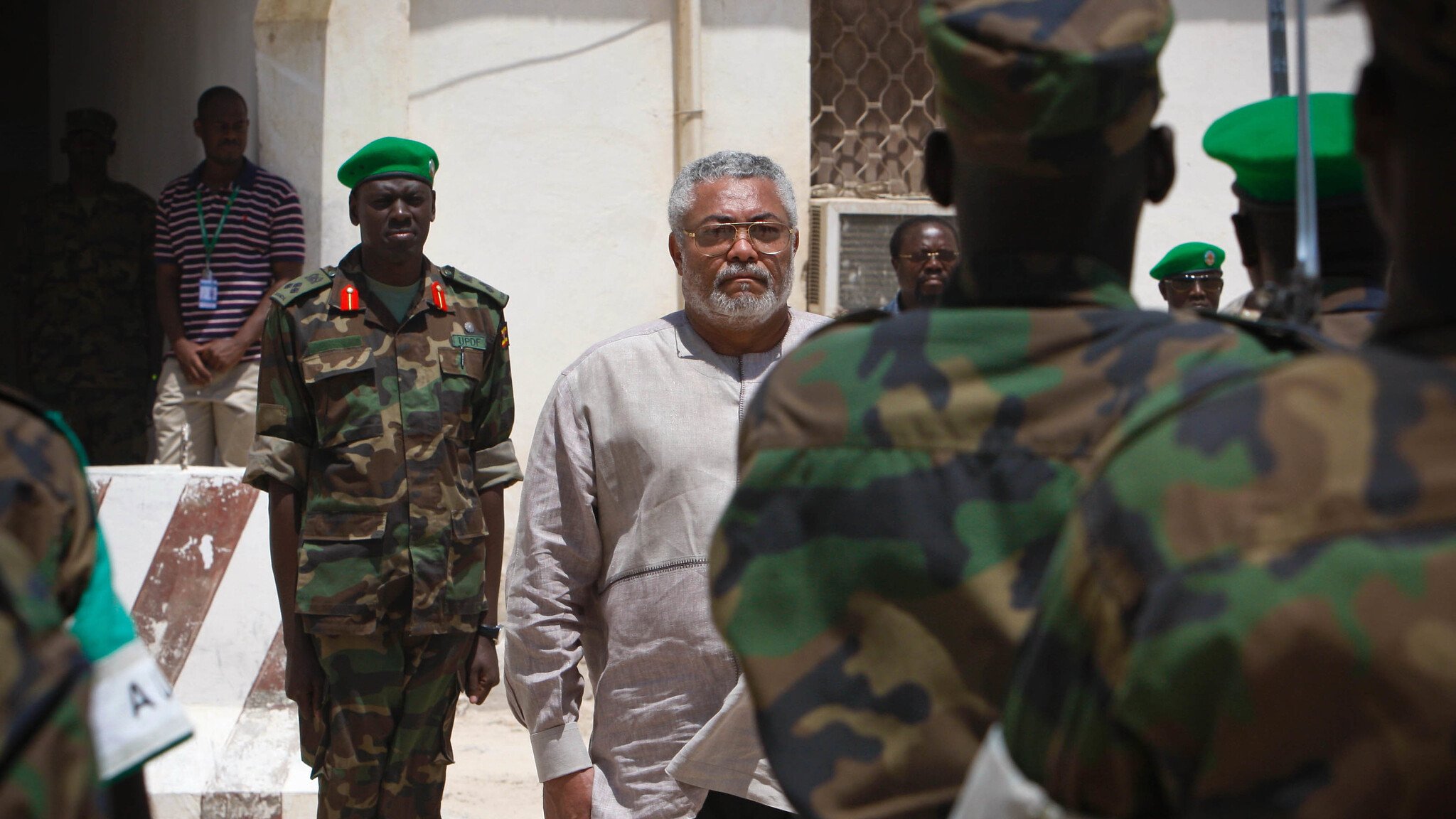 L'image montre un homme d'âge moyen au centre, avec des cheveux poivre et sel et une barbe grise. Il porte une chemise claire. Il semble sérieux et concentré, entouré de militaires en uniforme camouflage. Ces soldats, qui se tiennent en rangs, regardent vers lui. En arrière-plan, on peut voir un bâtiment aux murs clairs et un voisinage qui évoque un environnement militaire. L'atmosphère est formelle, soulignant l'importance de la scène.