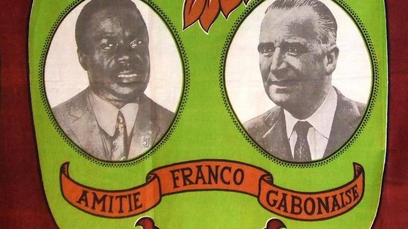 L'image présente un fond aux couleurs vives, dominé par des teintes vertes et marron. Au centre, deux portraits en noir et blanc sont encadrés. À gauche se trouve le portrait d'un homme à la peau noire, coiffé d'une chemise claire et affichant un regard sérieux. À droite, un homme à la peau claire, vêtu d'une chemise sombre, a une expression face à la caméra qui évoque la confiance. Entre les deux portraits, le mot "AMITIE" est inscrit en lettres majuscules, suivi de "FRANCO GABONAISE", soulignant un lien symbolique. Le motif de feuilles stylisées et d'ornements naturels en arrière-plan évoque un environnement tropical et chaleureux, symbolisant l'unité et la collaboration entre les cultures.