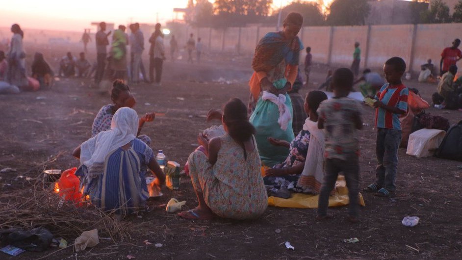 L'image montre une scène de vie en plein air, probablement à la tombée de la nuit. Au premier plan, plusieurs femmes et enfants sont assis sur le sol, entourés de quelques objets personnels et de restes de nourriture. Les femmes, habillées de vêtements colorés, semblent préparer un repas autour d'un feu. En arrière-plan, la lumière du crépuscule illumine légèrement la scène, et de nombreuses personnes peuvent être aperçues se déplaçant dans un environnement poussiéreux. L'atmosphère dégage à la fois une certaine convivialité et une sensation de précarité.