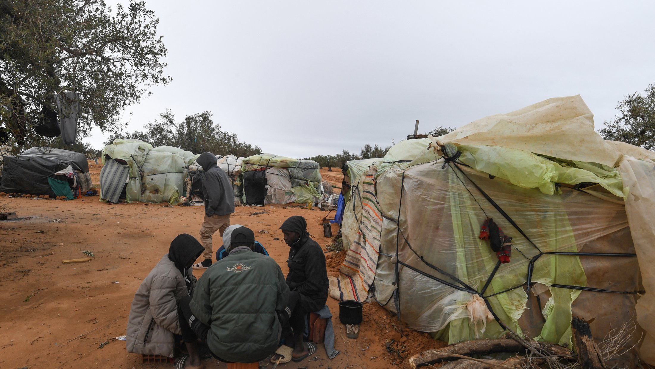 L'image montre un paysage d'abris précaires, construits avec des matériaux comme du plastique et des bâches, disposés sur un terrain sablonneux. Au premier plan, un groupe de personnes est assis en cercle, partagé entre une conversation ou une activité commune. Les abris semblent rudimentaires, entourés d'un environnement naturel avec quelques arbres épars. L'atmosphère est grise, suggérant un temps maussade. Skies sombres et nuageux donnent une impression de froid et de dureté.