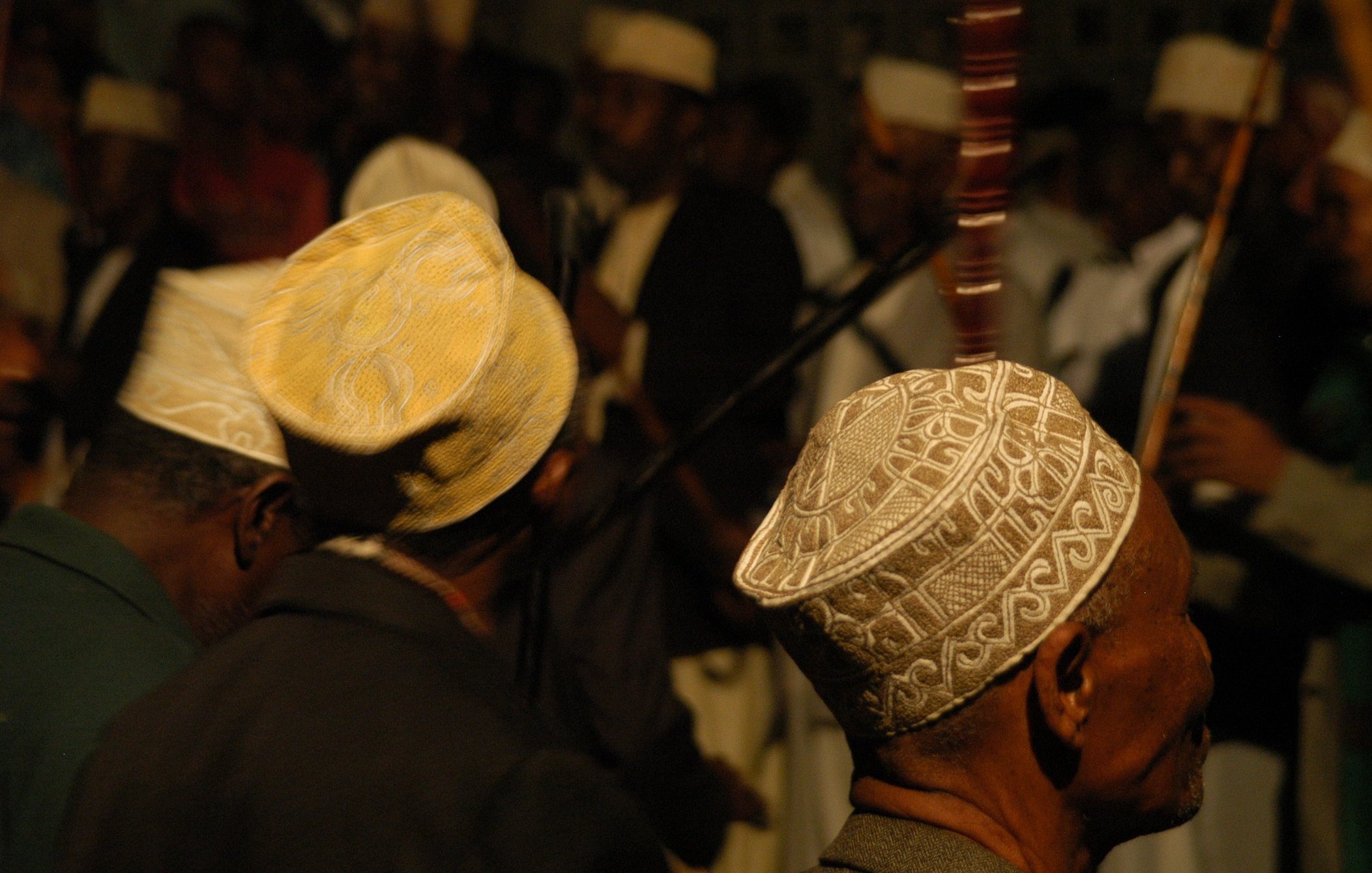 L'image présente un groupe de personnes, principalement des hommes, de profil. Ils portent des chapeaux traditionnels, souvent ornés de motifs délicats, qui varient en couleurs et en designs. En arrière-plan, on peut distinguer d'autres figures et instruments de musique, suggérant une ambiance festive ou rituelle. La lumière est tamisée, créant une atmosphère intime. Les visages des hommes sont flous, mais leur posture et leurs chapeaux ajoutent une touche culturelle à la scène.