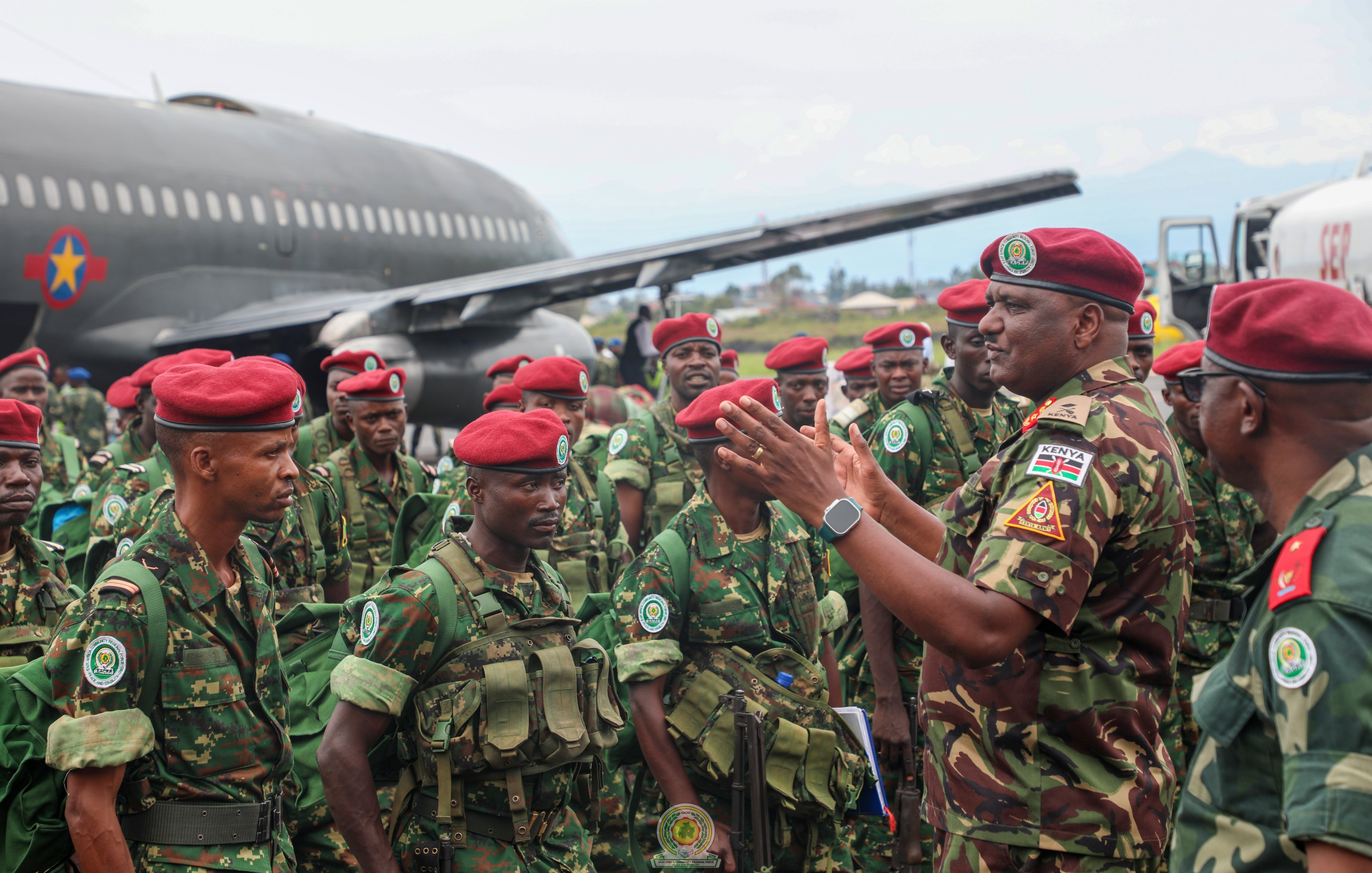 L'image montre un groupe de soldats en uniforme militaire, réunis près d'un gros avion de transport. Les soldats portent des casques et des berets rouges, et ils sont habillés de camouflages verts. L'un des officiers, qui semble donner des instructions ou des encouragements, se tient au centre, gesticulant avec les mains. En arrière-plan, on peut voir l'avion de taille importante, avec des couleurs de camouflage, sur un terrain d'aviation, tandis que le ciel est légèrement nuageux. L'atmosphère est sérieuse, témoignant d'une mobilisation militaire.