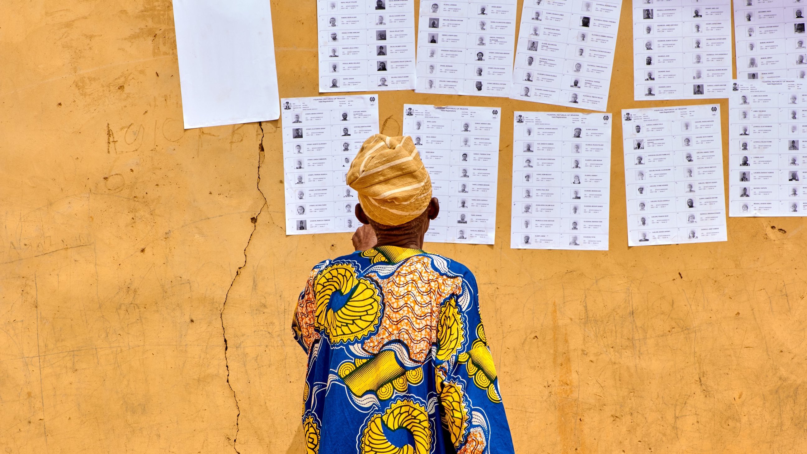 L'image montre un homme de dos, vêtu d'une robe colorée avec des motifs traditionnels, principalement en bleu et jaune. Il se tient devant un mur de couleur ocre, où sont affichés plusieurs documents. Ces documents ressemblent à des bulletins de vote ou des listes, alignés verticalement, avec des photographies et des noms inscrits. Le sol est légèrement fissuré, ajoutant une texture au fond de l'image. L'homme porte un turban beige, et son attention semble focalisée sur les informations affichées sur le mur.