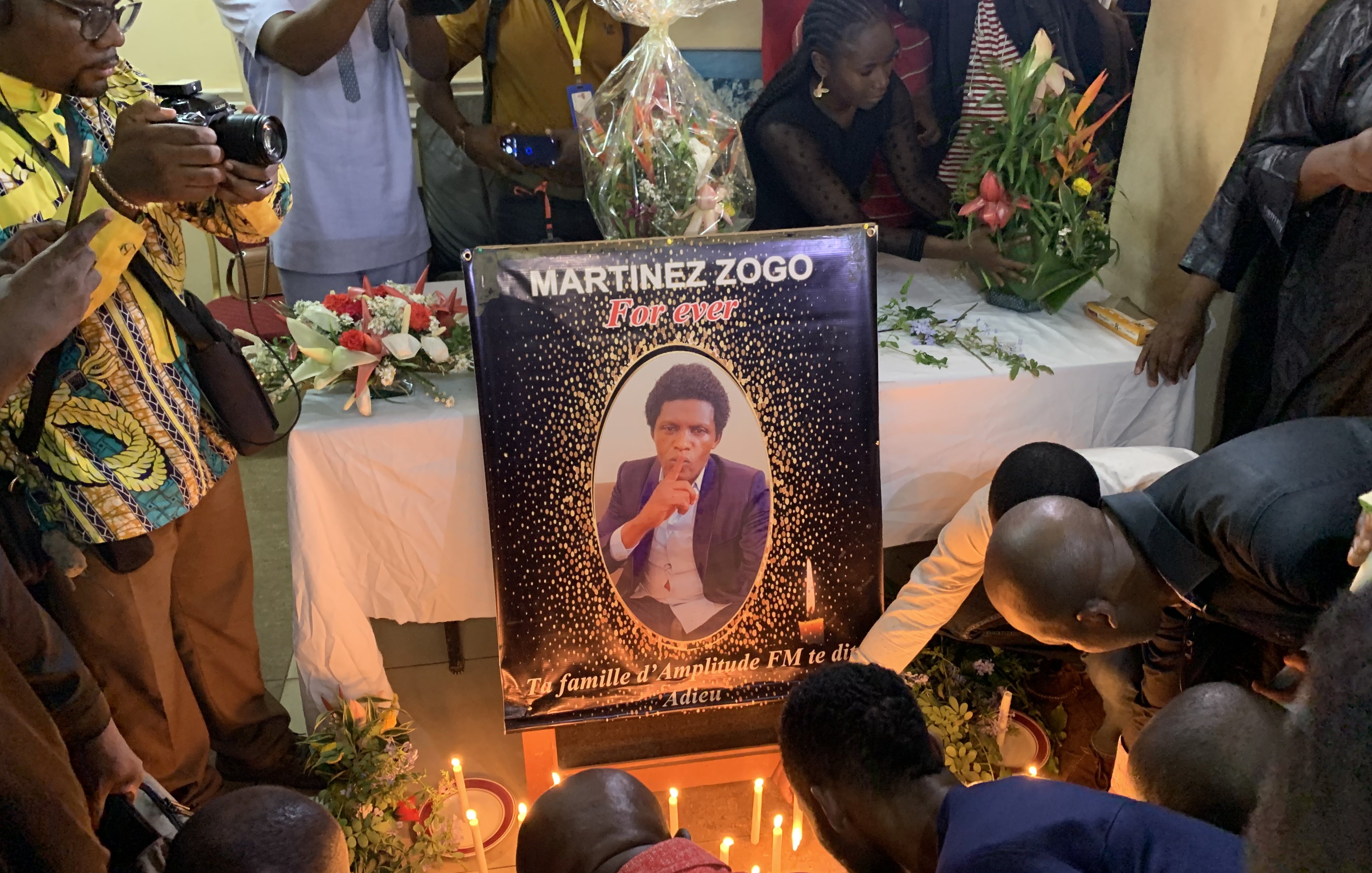 L'image dépeint une scène de recueillement. Au centre, il y a un grand poster représentant Martinez Zogo, encadré de fleurs et de bougies allumées. Le poster montre une photo de lui en train de fumer une cigarette, avec un regard pensif. Autour du poster, plusieurs personnes sont rassemblées, certaines s'agenouillant ou se penchant pour prier. L'atmosphère est solennelle, avec des expressions de tristesse sur les visages. Des fleurs et des bougies décorent la table sur laquelle le poster repose, ajoutant une touche de commémoration à cet hommage.