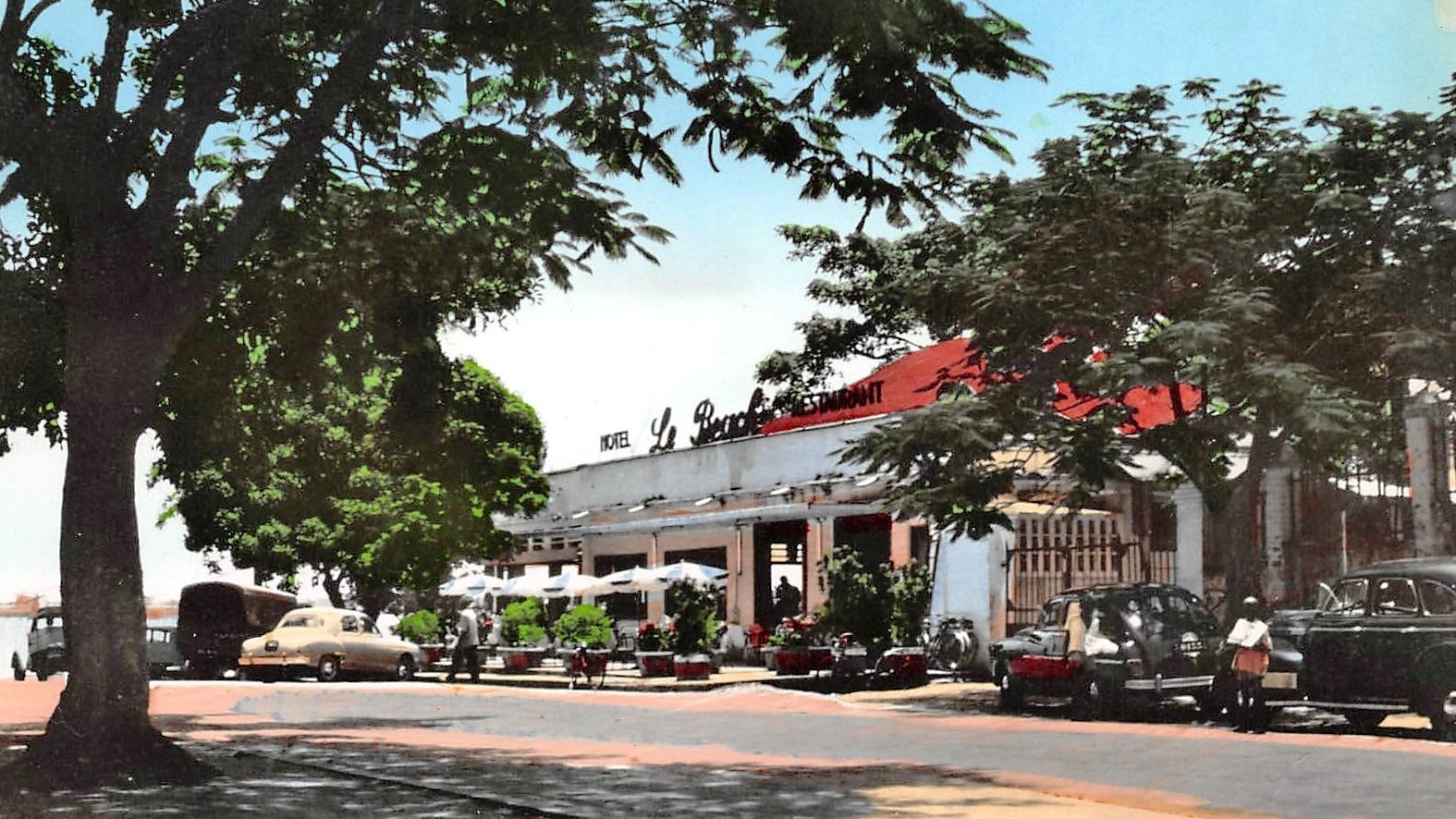 L'image montre une scène d'un restaurant, probablement située dans un quartier ensoleillé. À l'avant, on aperçoit une route bordée d'arbres dont les feuilles offrent un peu d'ombre. Le bâtiment principal, assez spacieux, possède un toit rouge et est marqué par l'inscription "Le Restaurant" en lettres blanches. Plusieurs voitures anciennes, de différentes couleurs, sont garées près de l'établissement. Des personnes flânent autour, créant une atmosphère de vie et de convivialité. En arrière-plan, le ciel est bleu, ce qui ajoute une touche de chaleur et de clarté à l'ensemble de l'image.