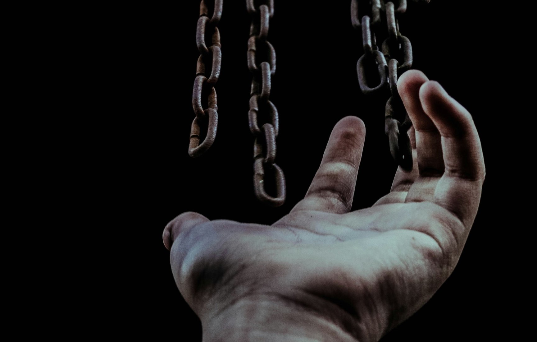 L'image montre une main tendue vers le haut, en direction de chaînes suspendues qui descendent. La main, légèrement ouverte, semble chercher à atteindre les chaînes, qui sont bien visibles sur un fond sombre. Les chaînes sont métalliques, avec une texture rustique, et elles évoquent une sensation d'emprisonnement ou de lutte. L'éclairage met en valeur les détails des chaînes et de la main, créant une atmosphère à la fois troublante et poignante. Cette composition suggère un thème de quête ou de désir de liberté.
