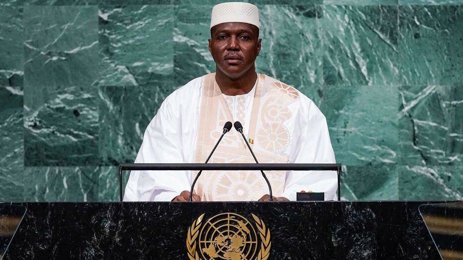 L'image montre un homme debout derrière un pupitre lors d'un discours. Il porte un costume traditionnel, probablement en blanc, avec un embellissement distinctif. Son teint est sombre et il a une expression sérieuse, indiquant qu'il est en train de s'adresser à un public. Le fond de l'image est constitué de panneaux de marbre vert, ce qui crée un contraste avec sa tenue. Le pupitre est orné du symbole des Nations Unies, représentant la mission internationale de paix et de coopération.