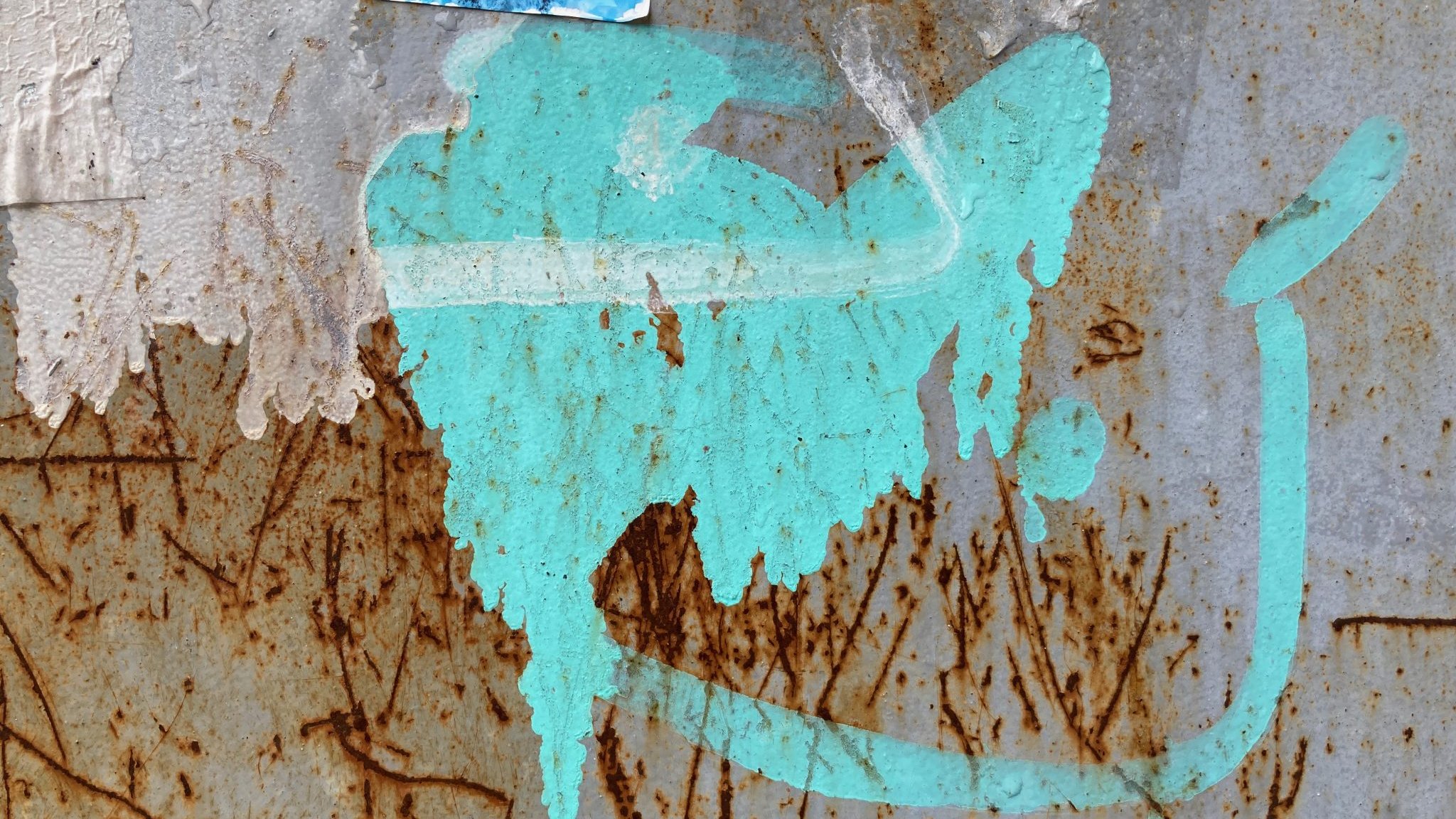 L'image présente une surface en métal, probablement rouillée, avec des marques et des textures variées. On y voit une tache de couleur turquoise qui semble avoir été projetée. Cette tache a une forme fluide et irrégulière, évoquant une sorte de mouvement ou de dynamisme. À proximité, on peut distinguer des traces brunes, résultant de la rouille, qui ajoutent un contraste marqué à la couleur vive. Le fond est texturé, avec des stries et des imperfections, suggérant l'âge et l'usure du matériau. L'ensemble crée une atmosphère à la fois urbaine et artistique.