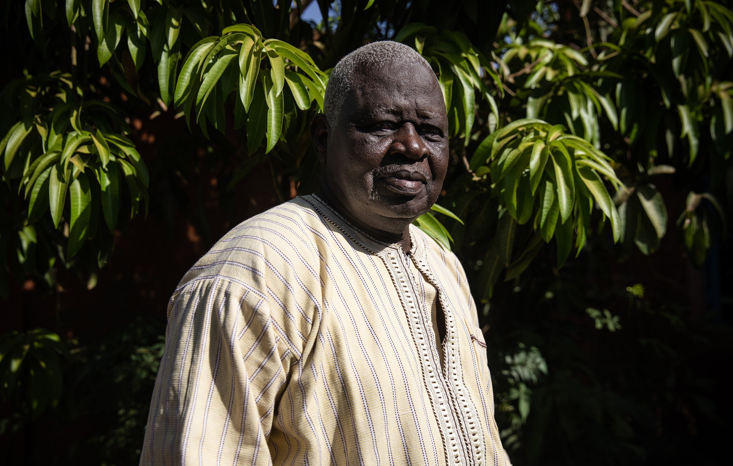 L'image présente un homme debout dans un environnement extérieur verdoyant. Il porte une tunique ample de couleur claire, ornée de motifs discrets, qui témoigne d'un style traditionnel. Son visage est marqué par des traits qui dégagent une certaine sagesse et sérénité. Les cheveux de l'homme sont grisonnants, ajoutant à son apparence mature. En arrière-plan, on aperçoit un feuillage dense et luxuriant, créant un contraste avec sa tenue. La lumière naturelle met en valeur les détails de son visage et de son vêtement, tout en évoquant une atmosphère paisible et accueillante.
