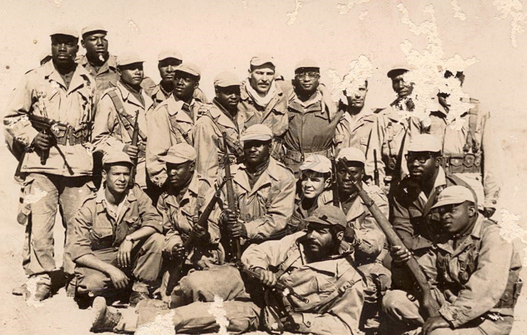 Cette image montre un groupe de soldats, probablement dans un contexte historique. Ils sont vêtus d'uniformes militaires, la plupart portant des chapeaux ou casquettes. Certains d'entre eux tiennent des armes, et l'ambiance générale dégage un sentiment de camaraderie et de force. Les soldats semblent être regroupés, avec une variation dans leurs postures : certains se tiennent debout tandis que d'autres sont accroupis ou assis. Le fond de la photo est déteint, ce qui suggère qu'il pourrait s'agir d'une image ancienne. L'ensemble de cette scène évoque un moment de solidarité et de détermination au sein du groupe.