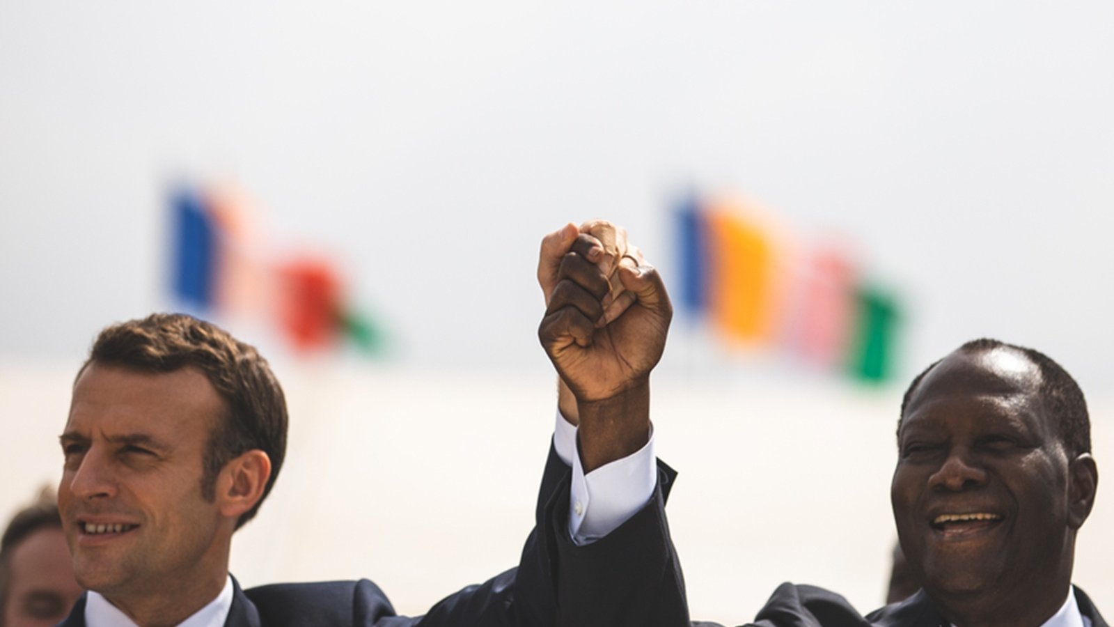 Imaginez une scène marquée par un moment de camaraderie et d'unité. Deux hommes se tiennent côte à côte, levant la main droite en l'air, leurs poings serrés, symbolisant la force et la solidarité. À gauche, on perçoit un homme avec une coupe de cheveux soignée, vêtu d'un costume sombre, tandis que l'homme à droite, également en costume, affiche un grand sourire chaleureux. En arrière-plan, plusieurs drapeaux flottent doucement au vent, représentant différentes nations. L'atmosphère est festive et pleine d'espoir.