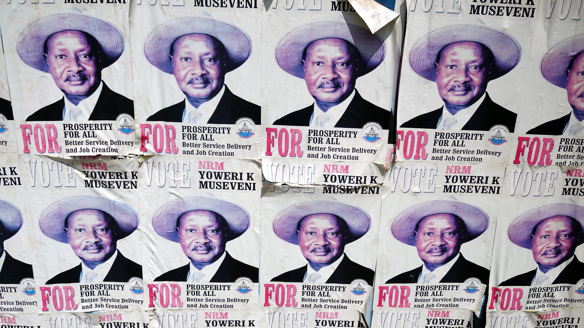 L'image montre un collage de plusieurs affiches sur un mur. Chaque affiche présente le même portrait d'un homme portant un chapeau de paille. Il a un grand sourire et affiche une expression confiante. Sous son image, on peut lire des textes en lettres majuscules qui soulignent des mots comme "PROSPERITY" (prospérité) et "FOR" (pour). Les affiches semblent faire partie d'une campagne politique, faisant appel au soutien des électeurs. Le fond est en papier décoloré et usé, indiquant que ces affiches ont été exposées pendant un certain temps.