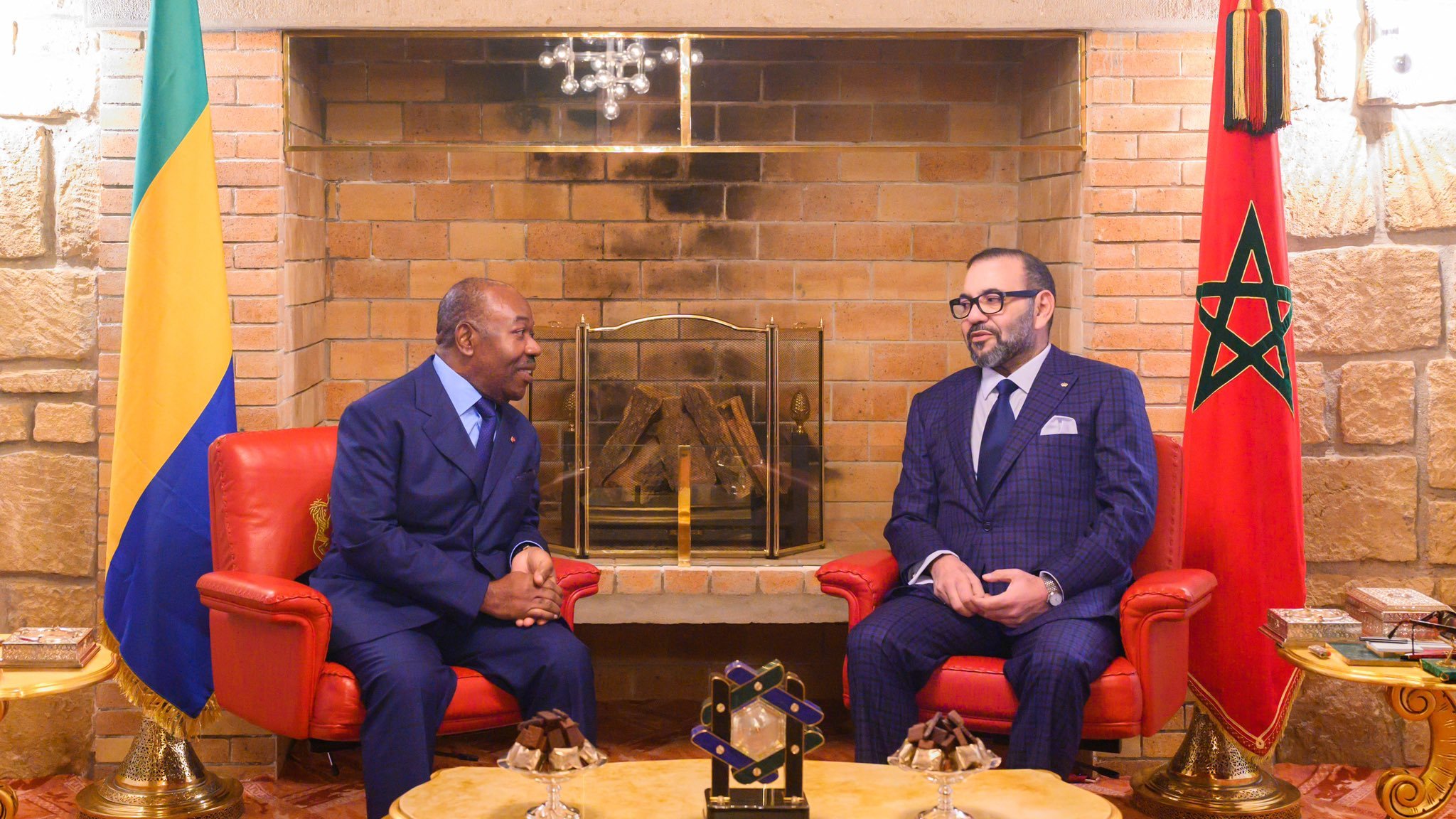 L'image montre deux hommes assis face à face dans un salon chaleureux et élégant. À gauche, un homme en costume bleu, avec une cravate, est légèrement incliné vers l'avant, écoutant attentivement. À droite, un autre homme porte un costume à carreaux bleu foncé et une cravate assortie, affichant une expression sérieuse tout en participant à la conversation. Derrière eux, on peut voir un mur en briques claires et une cheminée décorative. Il y a également des éléments de décoration comme des fauteuils rouges et un tableau avec un drapeau marocain sur le côté droit. Une lumière douce illumine la pièce, créant une atmosphère conviviale.