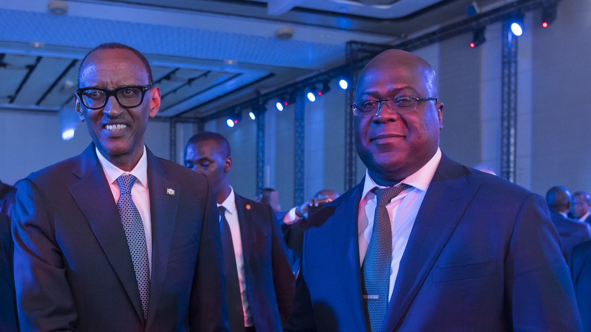 L'image montre deux hommes en costume qui se tiennent debout, se serrant la main dans un contexte de réunion ou d'événement officiel. L'un porte des lunettes et semble d'origine rwandaise, tandis que l'autre est un homme robuste, légèrement plus âgé, probablement d'origine congolaise. Ils affichent des expressions souriantes et amicales, dans une ambiance éclairée par des lumières bleues créant une atmosphère conviviale. En arrière-plan, on peut entrevoir d'autres personnes présentes à l'événement.