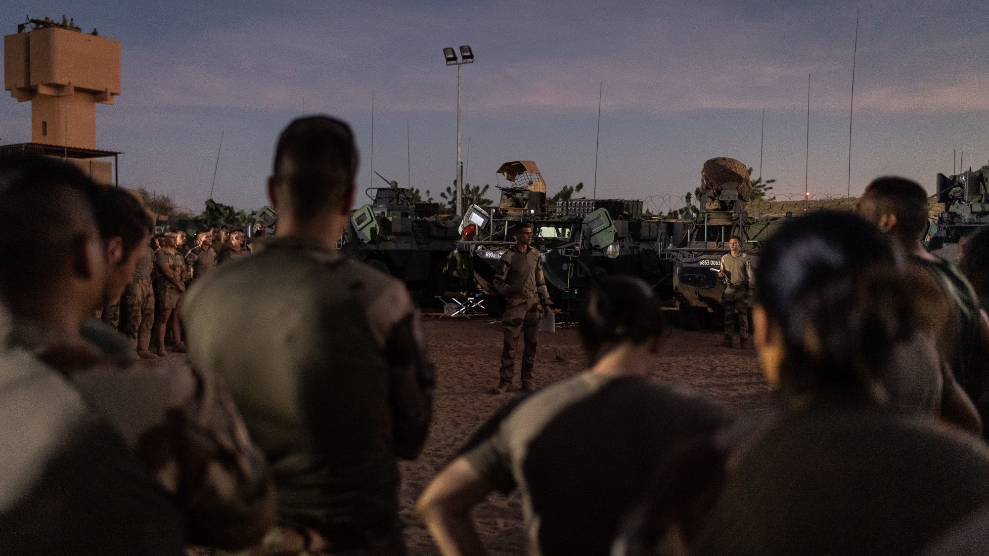 L'image montre un groupe de soldats rassemblés, attentifs, dans un environnement désertique au crépuscule. Ils sont partiellement de dos, ce qui met en avant leur tenue militaire. À l'arrière-plan, plusieurs véhicules militaires sont visibles, équipés d'armements. Le ciel est teinté de nuances douces, marquant la transition entre le jour et la nuit. L'atmosphère semble concentrée, suggérant une préparation ou un briefing important pour les soldats sur le terrain.