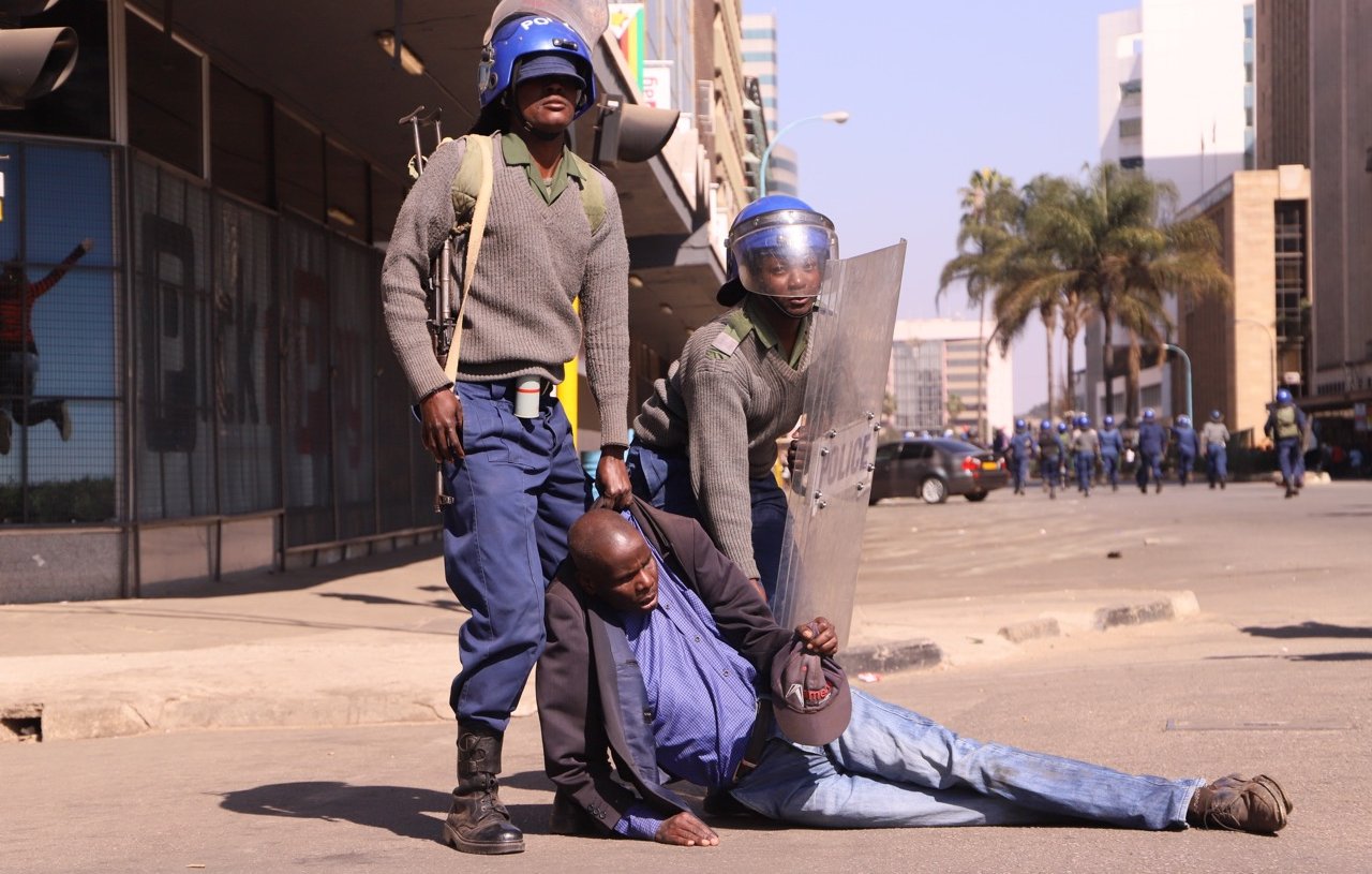 L'image montre une scène de tension dans un environnement urbain. Deux policiers, en uniforme et portant des casques de protection, semblent maîtriser un homme qui est au sol. L'homme est vêtu d'une chemise foncée et d'une veste. Il semble être en difficulté, soutenu par l'un des policiers, tandis que l'autre se tient à ses côtés avec un bouclier. En arrière-plan, on peut apercevoir des bâtiments modernes et quelques palmiers, indiquant que la scène se déroule dans une ville. L'atmosphère générale semble tendue, évoquant un moment de confrontation.