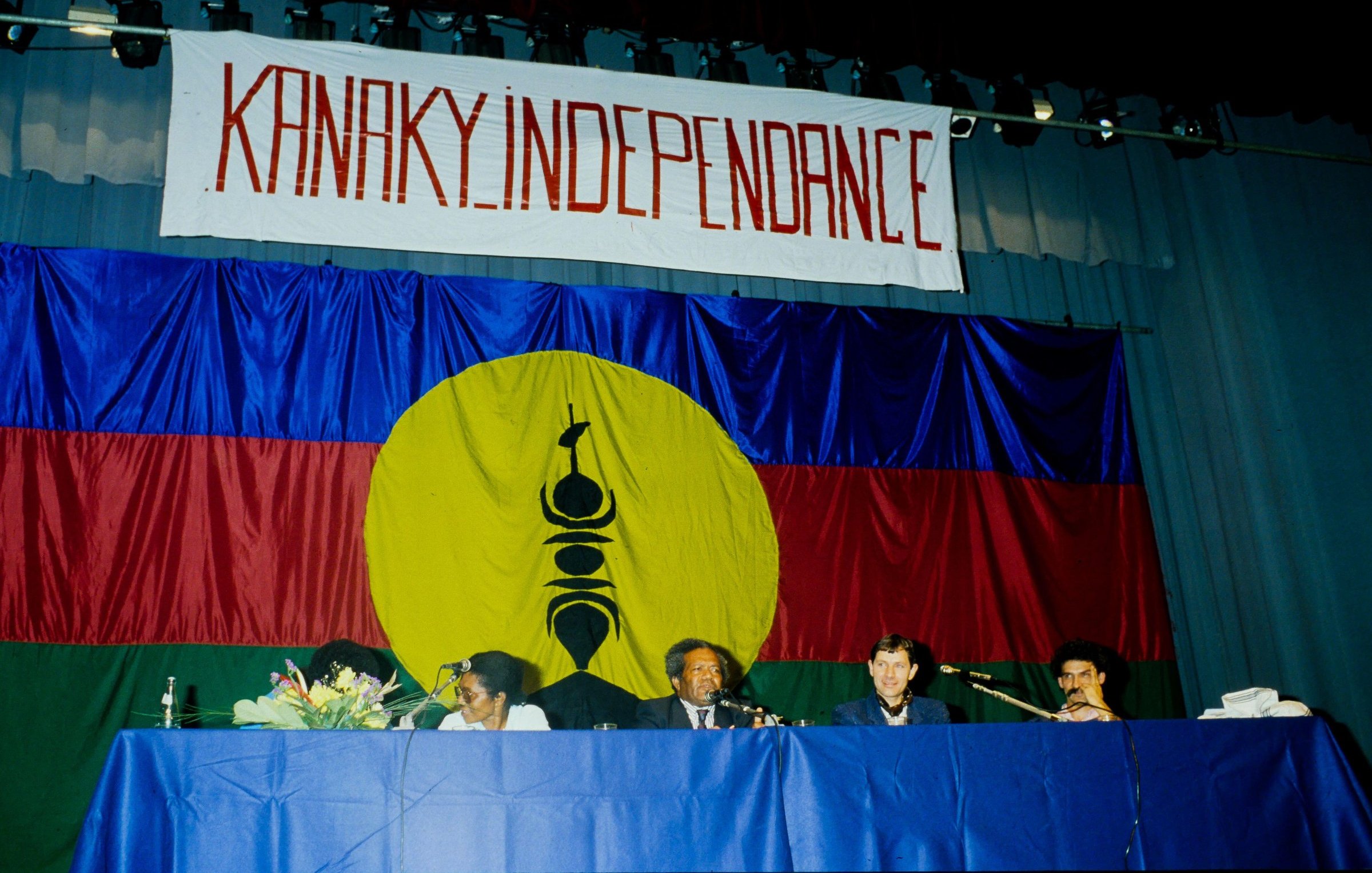 L'image présente une scène de réunion ou de conférence, où des personnes sont assises derrière une table. En arrière-plan, il y a un grand drapeau avec des couleurs vives : un rouge et un bleu, et un cercle jaune au centre. Dans ce cercle, on peut distinguer un symbole noir, précisant ainsi l'identité culturelle. Au-dessus de la scène, une banderole arbore le mot "KANAKY INDEPENDANCE" écrit en lettres rouges, exprimant un message fort lié à l'indépendance. Les participants semblent engagés et sont en discussion, créant une atmosphère de détermination et de solidarité.