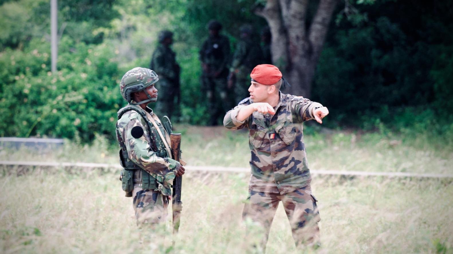L'image montre deux militaires en discussion dans un environnement extérieur. À gauche, un soldat vêtu d'un uniforme camouflage, portant un casque et tenant un fusil. À droite, un autre militaire, qui porte une tenue camouflage et un béret rouge. Il semble gesticuler en indiquant une direction. En arrière-plan, on aperçoit d'autres soldats flous, ce qui suggère qu'ils sont dans une zone d'entraînement ou opérationnelle, entourée de végétation et d'arbres. L'ambiance est sérieuse et dénote une situation de coordination ou de briefing entre les membres de l'armée.