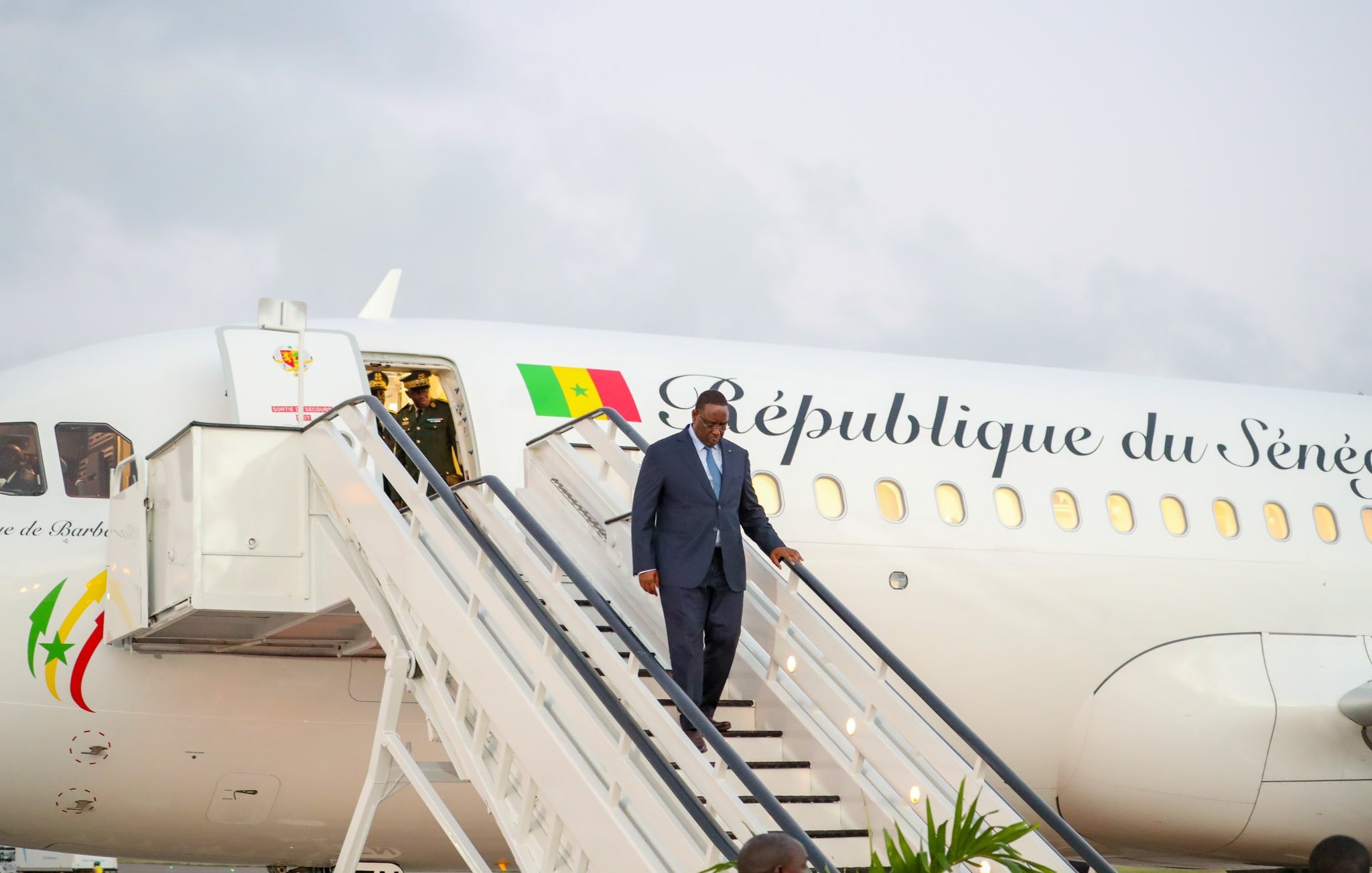 L'image montre un homme en costume sombre descendant d'un avion. L'avion, aux couleurs blanches et avec l'inscription "République du Sénégal" sur le fuselage, est garé sur une piste. À l'arrière-plan, le ciel est légèrement nuageux. Le personnage semble marcher avec assurance, et des escaliers mènent directement à la porte de l'avion. Au sol, on peut voir quelques plantes, suggérant un environnement accueillant.