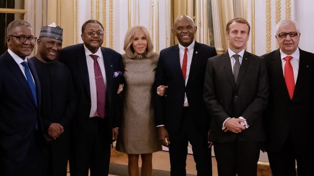 L'image montre un groupe de sept personnes se tenant debout dans une salle élégante, avec des murs clairs et des rideaux drapés. Au centre, une femme souriante porte une robe brillante et se tient entourée de six hommes en costume. Chacun d'eux a un regard chaleureux et engageant. Les hommes varient en teinte de peau et en style vestimentaire, certains portant des cravates, d'autres des vêtements traditionnels. L'ambiance de l'image dégage une atmosphère professionnelle et amicale, suggérant un moment de convivialité ou une rencontre importante.