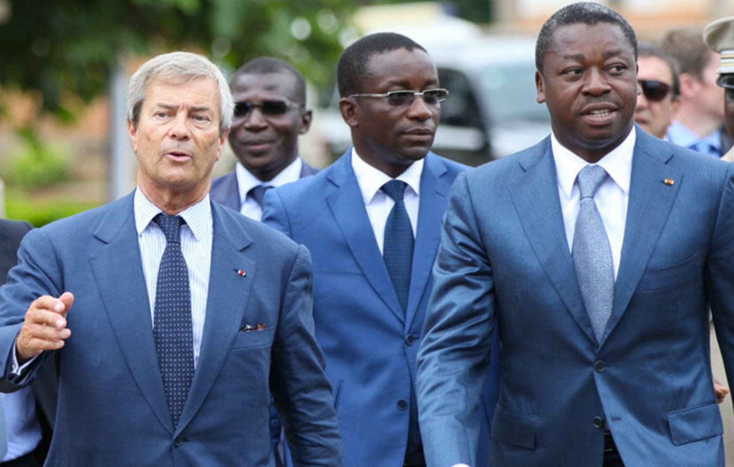 L'image montre un groupe d'hommes marchant ensemble. Le premier homme, à gauche, porte un costume bleu clair et une cravate à rayures. Il semble engagé dans la conversation. Le deuxième homme, au centre, porte aussi un costume bleu, avec une chemise blanche. Il a un regard sérieux et écoute attentivement. Le troisième homme, à droite, est vêtu d'un costume bleu foncé et marche avec assurance, affichant un léger sourire. En arrière-plan, on aperçoit d'autres individus et un environnement qui suggère un événement ou une rencontre formelle. Les hommes semblent dans un contexte professionnel.