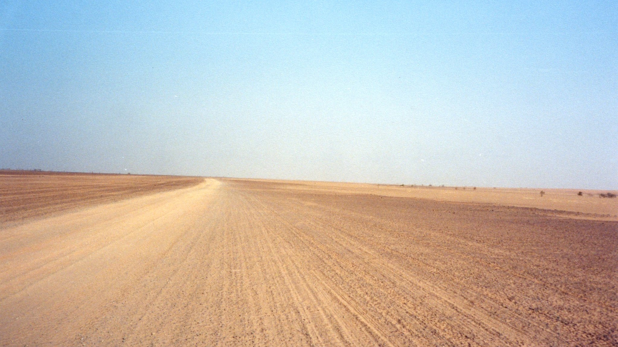 Imaginez un vaste paysage désertique, s'étendant à perte de vue sous un ciel bleu clair. Le sol est constitué de sable et de terre, créant une texture rugueuse sous vos pieds. La route est légèrement sinueuse, formant des lignes qui se fondent avec l'horizon. À première vue, tout semble calme et silencieux, avec peu de perturbations, excepté pour le vent. Sur les bords de ce paysage, on peut percevoir des nuances jaune et beige, typiques des déserts. L'atmosphère est sereine, presque méditative, laissant une impression de solitude face à l'immensité de la nature.