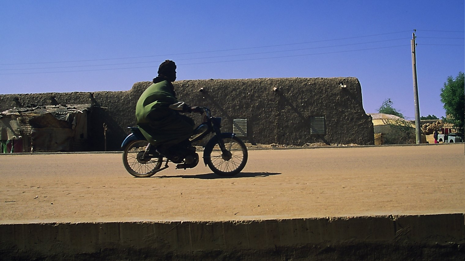 L'image montre un homme en train de rouler à bicyclette sur une route en terre. Il porte une tenue traditionnelle de couleur verte, qui contraste avec le décor. En arrière-plan, on peut apercevoir des bâtiments en adobe, typiques des régions désertiques, avec des murs de terre et des fenêtres. Le ciel est d'un bleu éclatant, sans nuages, et l'atmosphère générale est calme, suggérant une journée ensoleillée. La lumière du soleil éclaire la scène, mettant en valeur les textures du sol et des constructions.