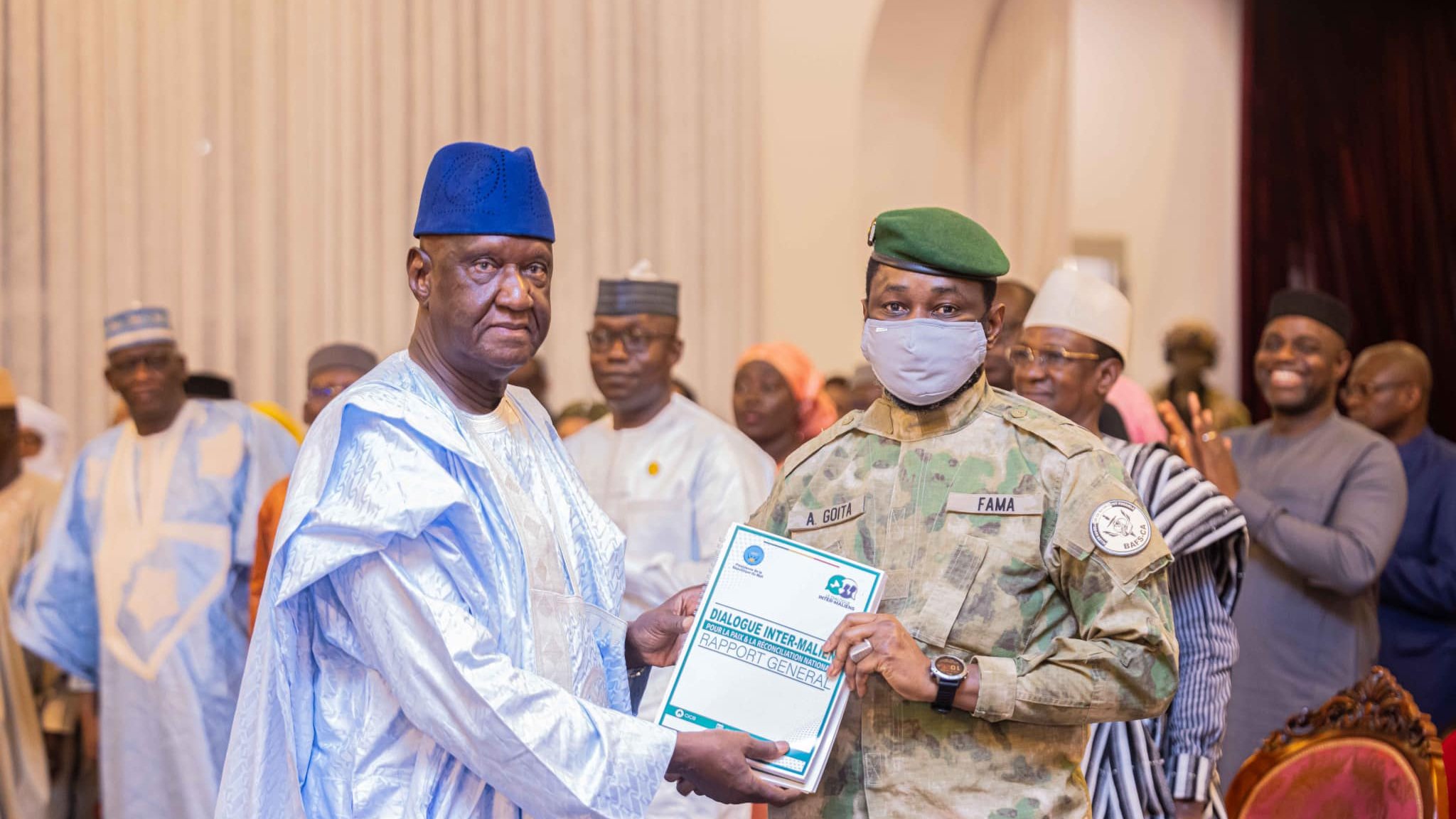 L'image montre un événement officiel ou une cérémonie de remise de prix. Au premier plan, un homme portant une tenue traditionnelle, composée d'une longue robe et d'un chapeau bleu, reçoit un document d'un officier militaire. Ce dernier est vêtu d'un uniforme camouflé et porte un masque gris. En arrière-plan, plusieurs personnes en tenue variée assistent à l'événement, certaines applaudissant, tandis que d'autres observent. L'environnement semble formel, avec des décorations adaptées à l'occasion.