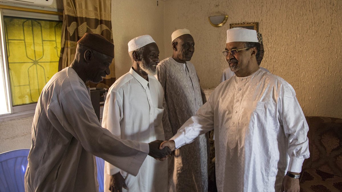 L'image montre un groupe de quatre hommes dans une pièce intérieure. Ils portent des vêtements traditionnels : des turbans et des vêtements clairs, souvent en coton. Un des hommes, sur la droite, est en train de serrer la main d'un autre homme, tandis que deux autres les regardent avec un air amical. La lumière entre par une fenêtre derrière eux, créant une atmosphère chaleureuse. Les murs sont texturés, avec un éclairage doux, et l'ensemble donne une impression de convivialité et de respect mutuel.