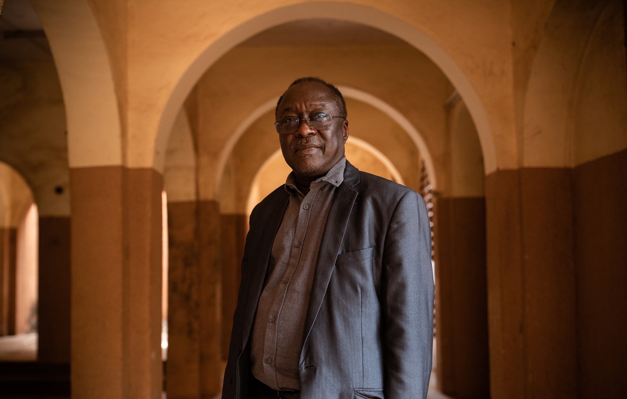 L'image montre un homme debout dans un espace intérieur, caractérisé par de grandes arcades. Les murs sont peints de couleurs chaudes, avec des tons ocre et beige. L'homme porte une veste sombre et une chemise grise, et il a l'air confiant. Son expression est sereine et il regarde légèrement vers la droite. L'arrière-plan présente des arches qui créent une atmosphère de profondeur et de perspective. La lumière semble douce et diffuse, ajoutant une ambiance chaleureuse à la scène.