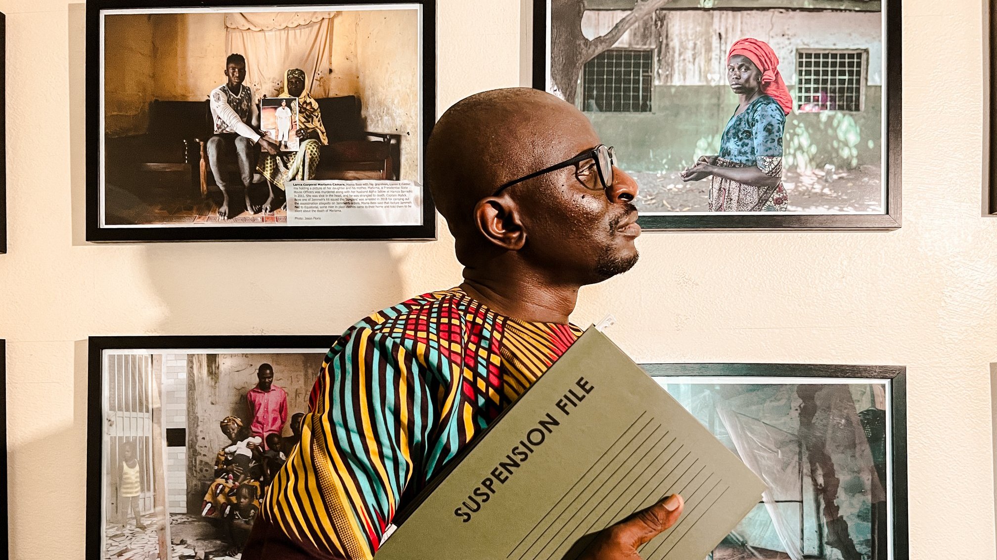 L'image montre un homme debout dans un espace d'exposition, entouré de plusieurs photographies accrochées au mur. Il porte une chemise colorée aux motifs vibrants, mêlant des teintes de jaune, rouge et vert. Dans ses bras, il tient un dossier intitulé "SUSPENSION FILE", ce qui suggère qu'il se prépare à discuter ou à présenter quelque chose d'important. L'homme se tourne légèrement sur le côté, regardant vers les photos, qui semblent raconter des histoires de vie, avec des portraits d'autres personnes. L'atmosphère de l'image est à la fois sérieuse et engagée, soulignant l'importance des histoires capturées dans ces œuvres visuelles.