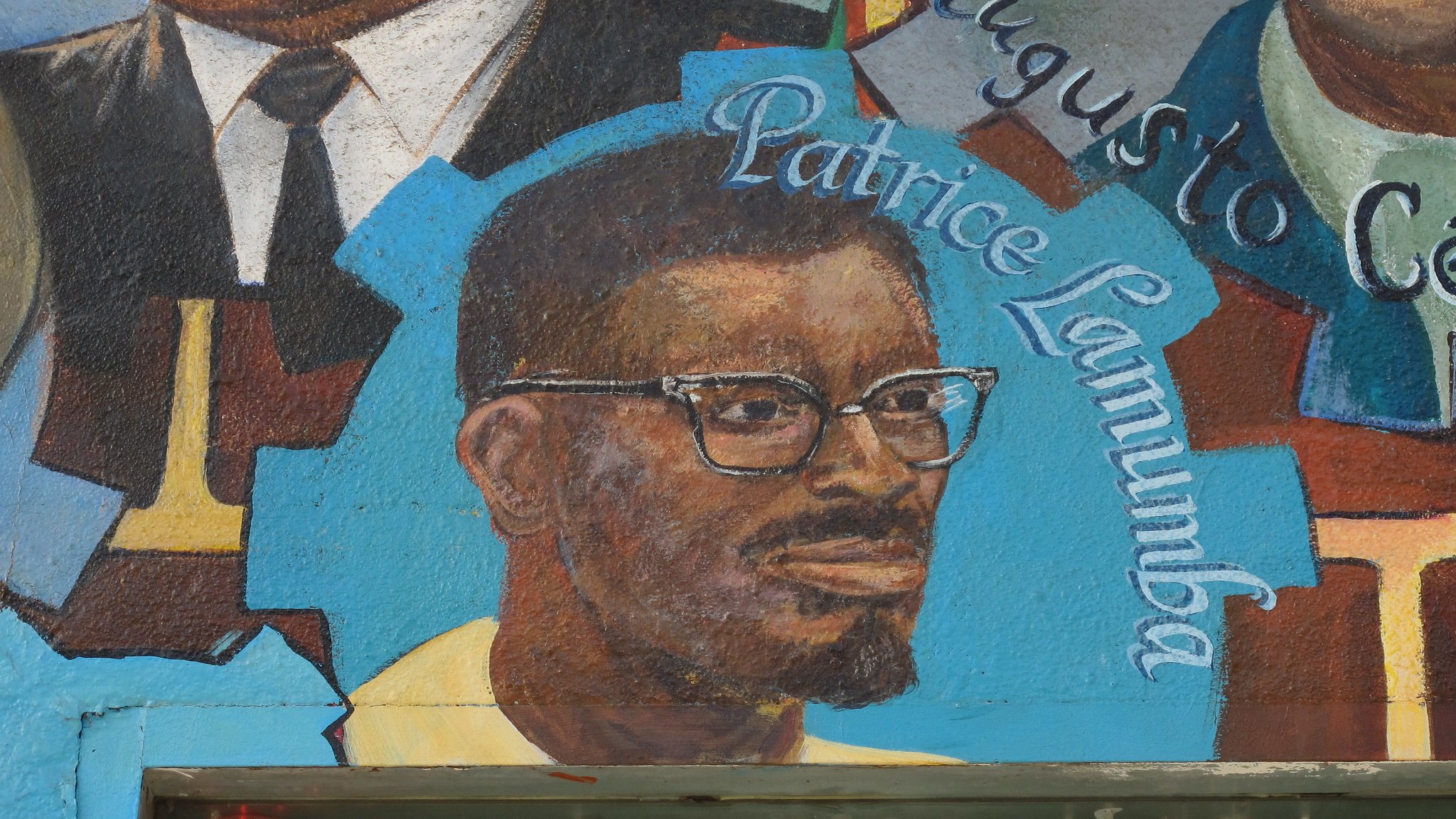 L'image semble représenter une fresque murale avec des portraits de différentes personnes. Au centre, on peut voir un homme portant des lunettes, avec une barbe et un sourire. Son nom, "Patrice Lumumba", est écrit en dessous de son portrait. Les autres éléments de la fresque peuvent inclure des symboles ou des motifs colorés, contribuant à une ambiance artistique et culturelle. La fresque pourrait célébrer des figures historiques ou politiques importantes.