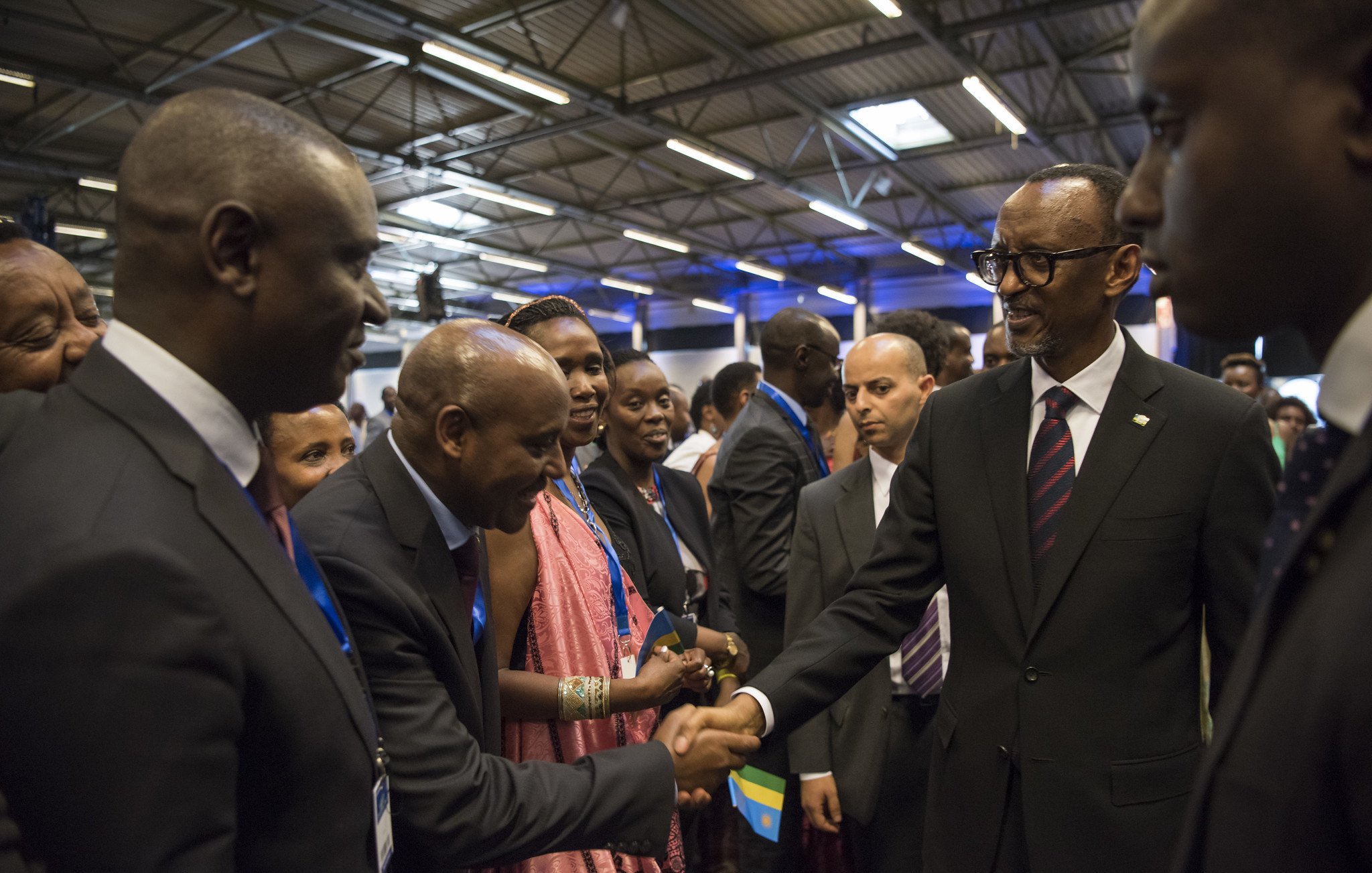Dans cette image, nous voyons un groupe de personnes rassemblées dans un espace intérieur, probablement lors d'un événement officiel ou d'une conférence. Parmi eux se trouve un homme en costume noir avec une cravate, qui semble être un dirigeant ou une personne importante. Il tend la main pour saluer un autre homme, également en costume. Les autres personnes présentes, principalement habillées formellement, regardent avec intérêt. L'atmosphère est conviviale et respectueuse, avec des sourires et des échanges amicaux. Les murs sont décorés de manière simple, et un éclairage doux illumine la scène.
