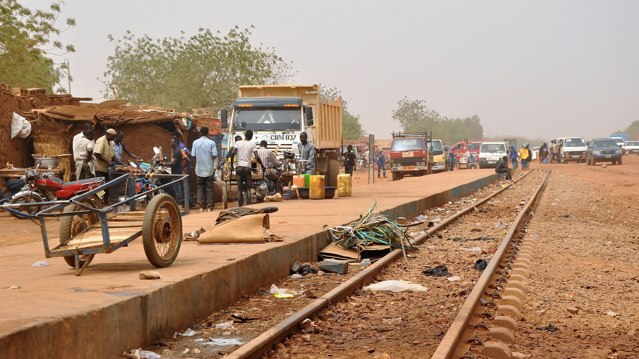 L'image dépeint une scène animée le long d'une voie ferrée. Sur la droite, des rails en métal s'étendent, entourés de poussière. À proximité, on aperçoit plusieurs véhicules, notamment des camions et des voitures, stationnés ou en mouvement. Des personnes se regroupent sur le côté, certains discutant, d'autres transportant des marchandises. Sur le sol, on peut distinguer divers objets laissés là, comme des déchets, des morceaux de métal et des câbles. À gauche, une petite charrette à deux roues est partiellement visible. En arrière-plan, la végétation est clairsemée, avec quelques arbres. Le ciel est d'un ton brumeux, suggérant une chaleur intense et un environnement rural. L'atmosphère générale est à la fois occupée et rustique, évoquant l'activité quotidienne dans une région où la vie s'organise autour des transports.