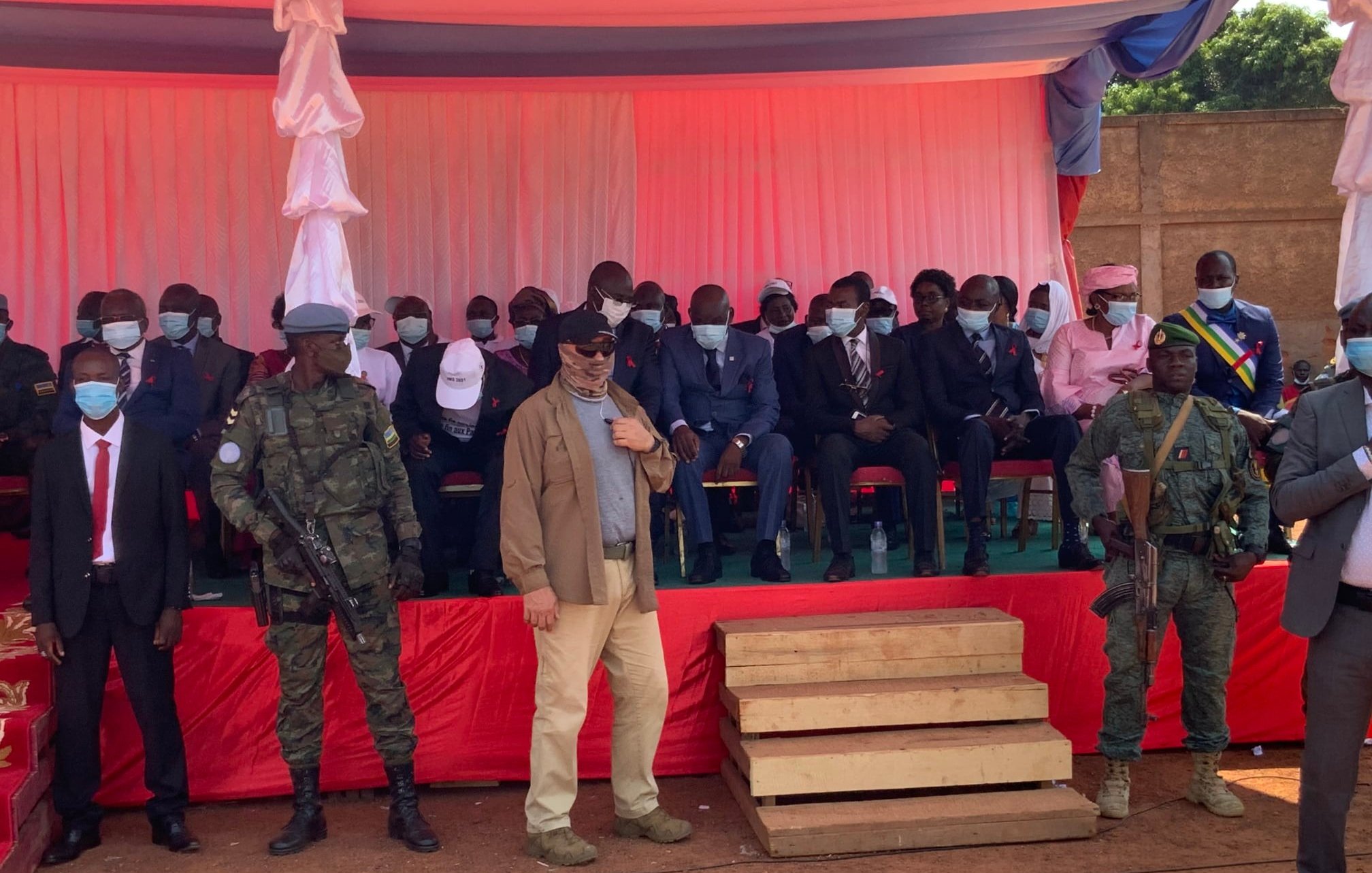L'image montre une scène d'événement en plein air, probablement une cérémonie officielle. Au premier plan, deux soldats en uniforme, l'un avec un casque et l'autre portant une veste militaire, se tiennent debout à côté de deux marches en bois. Derrière eux, un groupe de personnes est assis sur des chaises, en grande partie habillées de manière formelle, certaines portant des masques. Les hommes et les femmes sont alignés en rang, sur un fond de tissu rouge et blanc. L'ambiance semble solennelle, avec des drapeaux et des décorations typiques des événements institutionnels. Le décor est éclairé par une lumière naturelle, et l'atmosphère pourrait évoquer une cérémonie de discours ou une célébration publique.