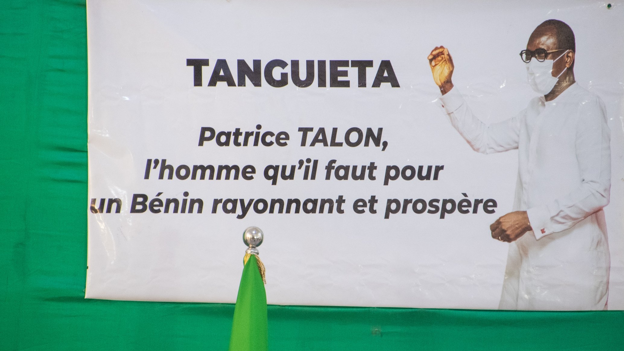 L'image présente une affiche accrochée sur un fond vert. Cette affiche met en avant le nom "TANGUIETA" en haut, suivi d'une mention concernant Patrice TALON. Le texte indique qu'il est "l'homme qu'il faut pour un Bénin rayonnant et prospère." On peut également voir un homme en tenue blanche, portant un masque. Il est positionné à droite de l'affiche, levant une main dans un geste de discours ou de présentation. À gauche de l'affiche, se trouve un drapeau vert qui se dresse à la verticale. L'ensemble véhicule un message d'espoir et de prospérité pour le pays.