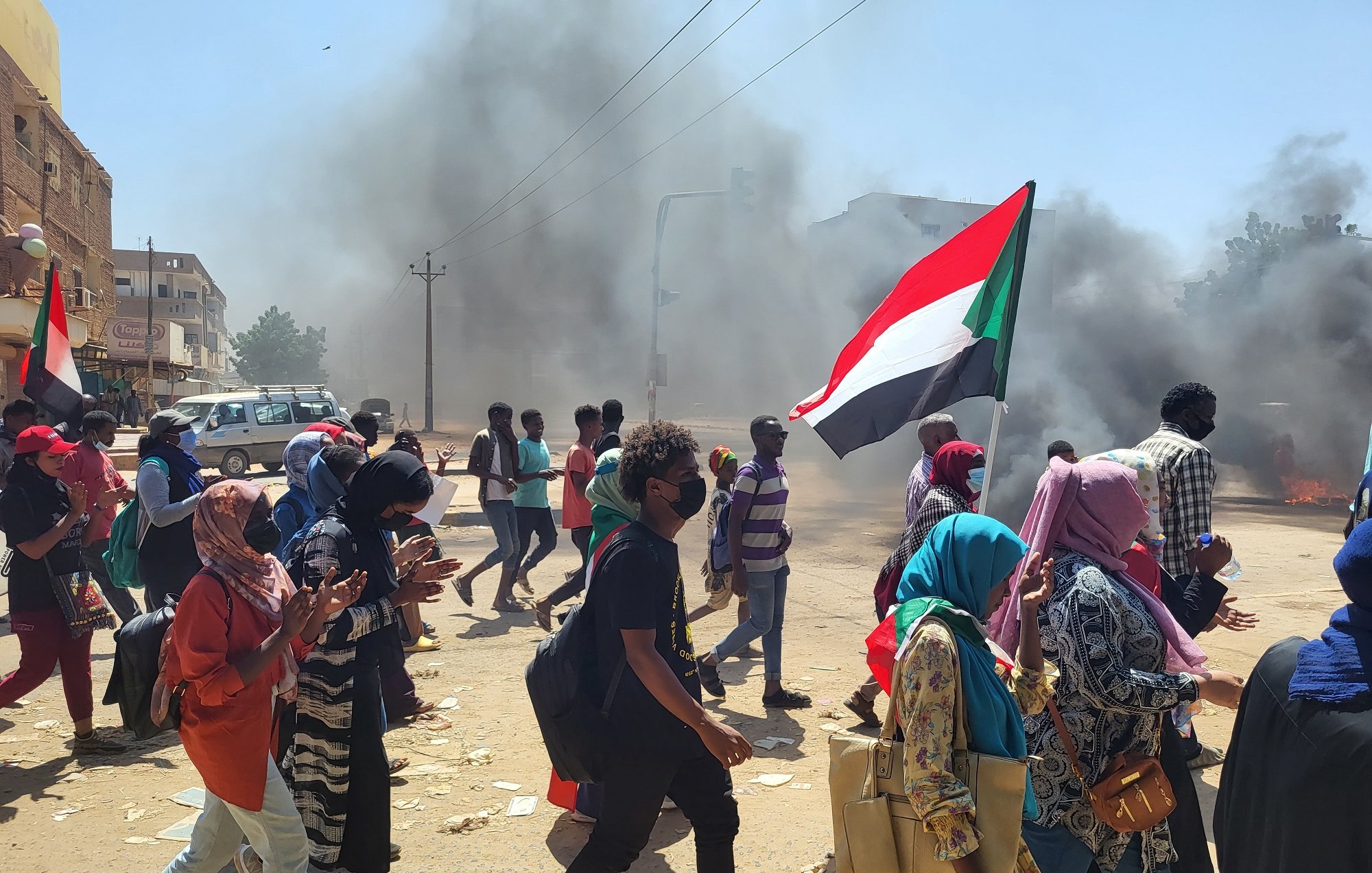 L'image montre une scène de manifestation animée. Des groupes de personnes marchent dans une rue, portant des drapeaux, dont un qui représente le Soudan. La foule paraît déterminée, et beaucoup de manifestants portent des masques, probablement en raison des conditions de pollution ou pour des raisons de sécurité. En arrière-plan, on peut voir des colonnes de fumée noire qui s'élèvent dans le ciel, créant une atmosphère tendue. Les manifestants semblent traverser une intersection, avec des véhicules garés sur le côté. L'atmosphère générale dégage une impression de forte mobilisation et de passion collective.