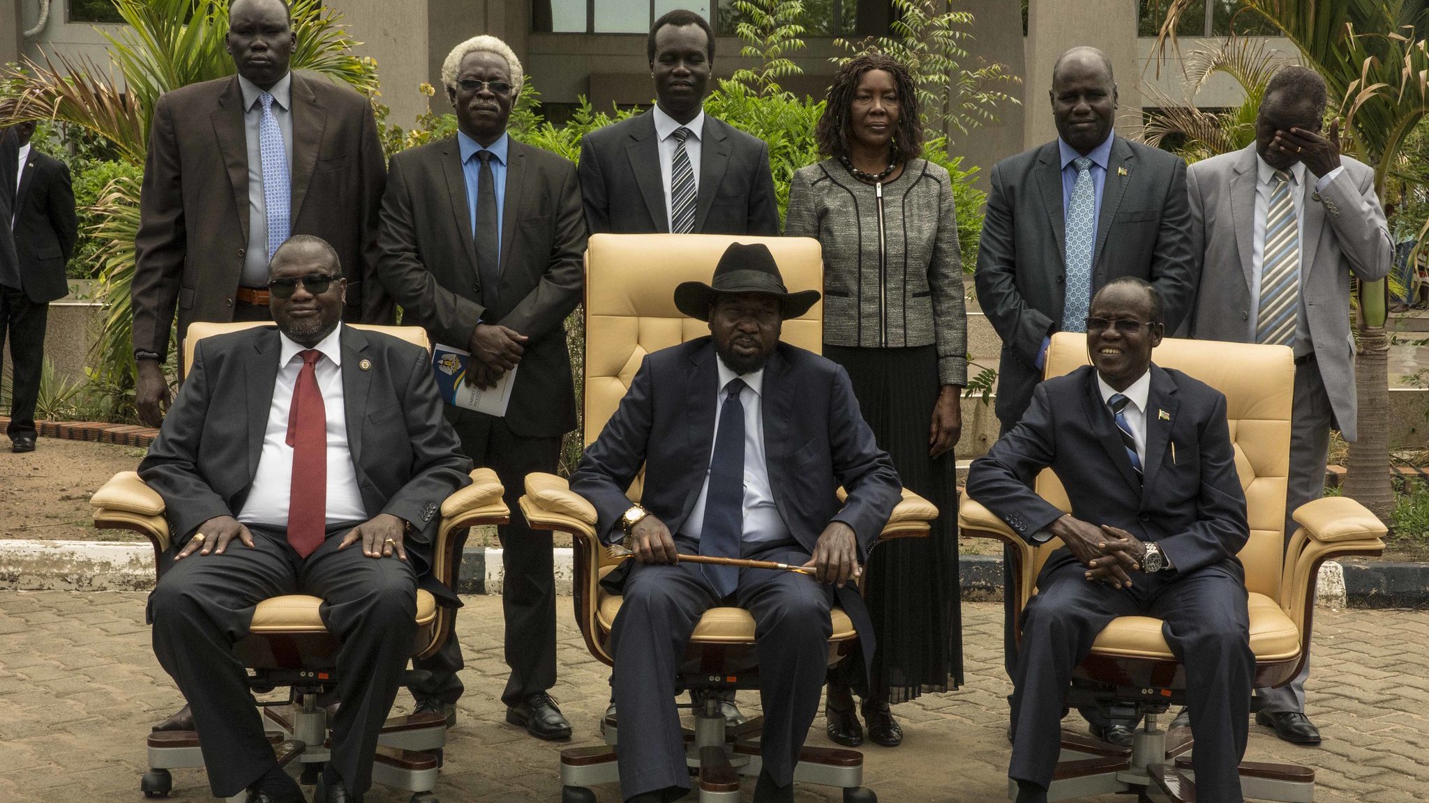 L'image présente un groupe de personnes posant ensemble à l'extérieur d'un bâtiment. Au centre, on voit trois hommes assis dans de grandes chaises confortables. Le premier homme à gauche porte des lunettes, un costume sombre et une cravate rouge. Le second, au centre, est vêtu d'un costume noir, d'une cravate et d'un chapeau noir, et semble être le leader du groupe. Le troisième homme, à droite, porte également un costume et une cravate. Derrière eux, plusieurs personnes se tiennent debout, toutes en tenue formelle. L'environnement est verdoyant, avec des plantes et un chemin en pavé. L'atmosphère semble sérieuse et officielle, suggérant une rencontre importante.
