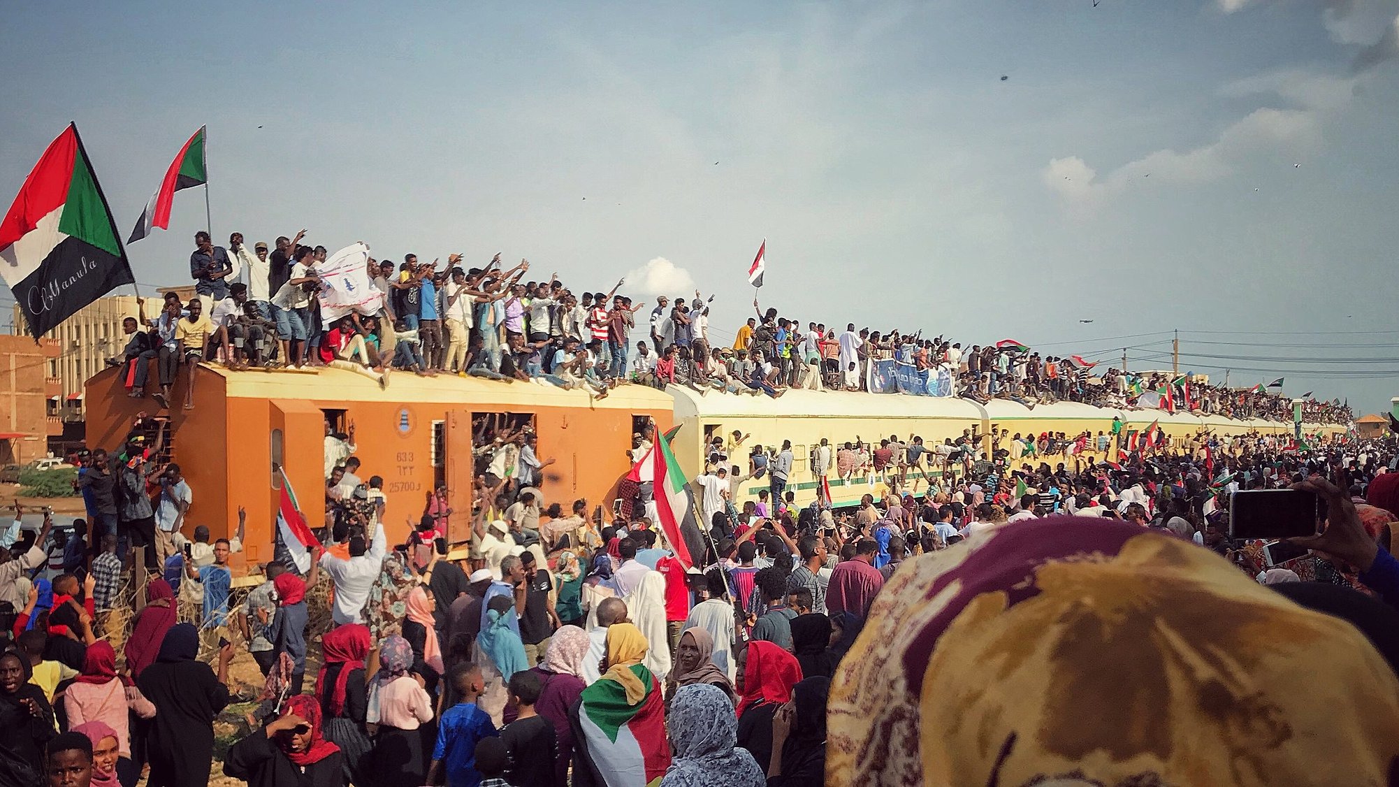 L'image montre une scène de grande effervescence, probablement lors d'un rassemblement ou d'une célébration. On peut voir une foule nombreuse, avec des gens debout sur un train, brandissant des drapeaux aux couleurs vives, en particulier du rouge, du noir et du vert. Les participants expriment leur enthousiasme et leur solidarité, certains levant les poings ou affichant des panneaux. La foule est diverse, incluant des personnes de tous âges, et il règne une atmosphère de ferveur collective, avec un ciel bleu parsemé de quelques nuages en arrière-plan. Cette image évoque des sentiments de joie, d'unité et de détermination.