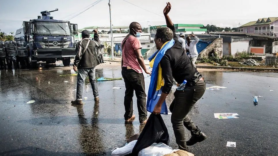 L'image montre une scène de manifestation dans une rue. Au premier plan, on peut voir des personnes en mouvement, certaines en train de gesticuler, tandis qu'une personne est à terre, apparemment blessée. L'un des manifestants porte un drapeau, probablement symbolique. En arrière-plan, il y a un véhicule de police, ce qui suggère une présence des forces de l'ordre. Le sol est couvert de débris, comme des affiches et des bouteilles, témoignant de la tension de la situation. L'ambiance semble intense et sérieuse, avec une impression de conflit.