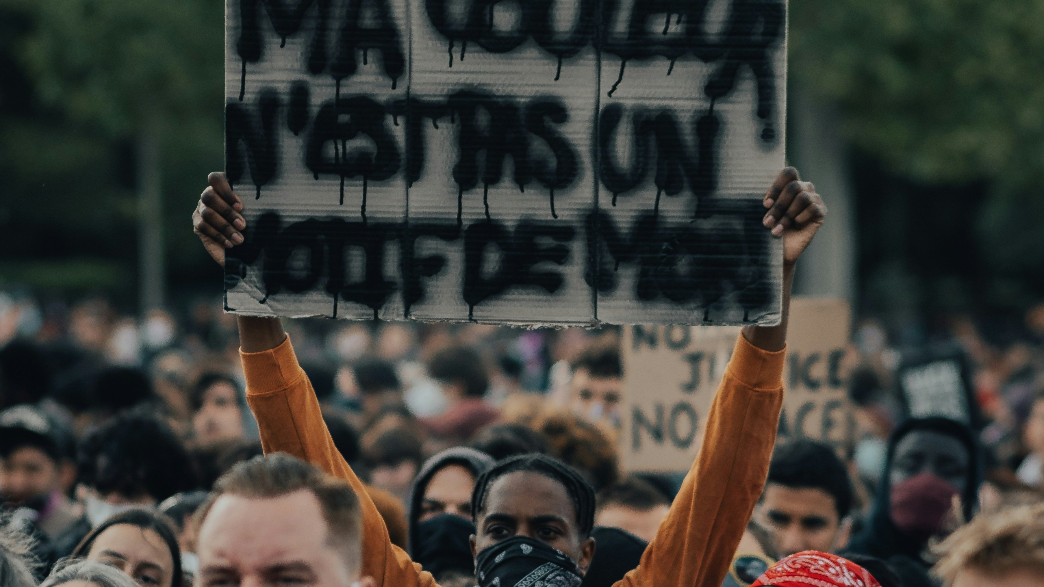 L'image montre une grande foule rassemblée pour une manifestation. Parmi les manifestants, une personne se distingue en tenant une pancarte sur laquelle est écrit "MA COULEUR N'EST PAS UN CRIME". Cette personne porte un masque, comme beaucoup d'autres dans la foule. Les visages autour sont sérieux et déterminés, reflétant un sentiment de solidarité et de revendication. On peut sentir l'intensité de l'événement grâce à l'énergie collective. En arrière-plan, des arbres indiquent un cadre naturel, ajoutant à l'atmosphère de mobilisation pacifique.