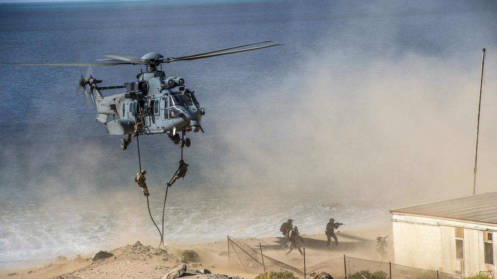 L'image montre un hélicoptère militaire en pleine action, en train de décoller ou de se poser près d'une côte. On peut entendre le bruit des pales qui tournent rapidement, créant un nuage de poussière et de sable autour de lui. À proximité, des soldats descendent en rappel, suspendus à une corde, tandis qu'ils avancent vers une zone de terrain difficile. En arrière-plan, on aperçoit la mer, calme, et une structure qui ressemble à un bâtiment près de la plage. L'atmosphère générale dégage un sentiment d'urgence et d'opération militaire.