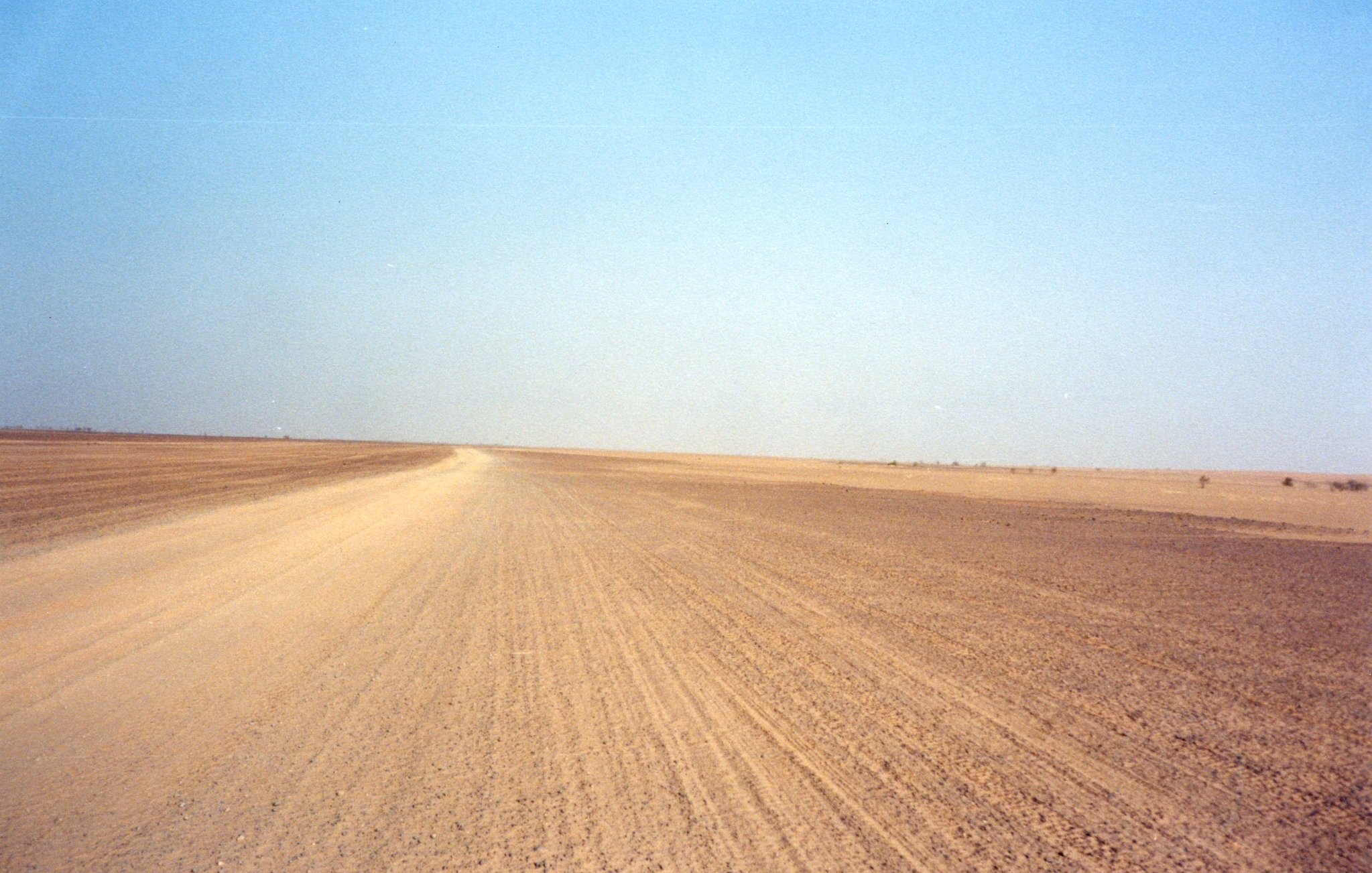 Imaginez un vaste paysage désertique, s'étendant à perte de vue sous un ciel bleu clair. Le sol est constitué de sable et de terre, créant une texture rugueuse sous vos pieds. La route est légèrement sinueuse, formant des lignes qui se fondent avec l'horizon. À première vue, tout semble calme et silencieux, avec peu de perturbations, excepté pour le vent. Sur les bords de ce paysage, on peut percevoir des nuances jaune et beige, typiques des déserts. L'atmosphère est sereine, presque méditative, laissant une impression de solitude face à l'immensité de la nature.
